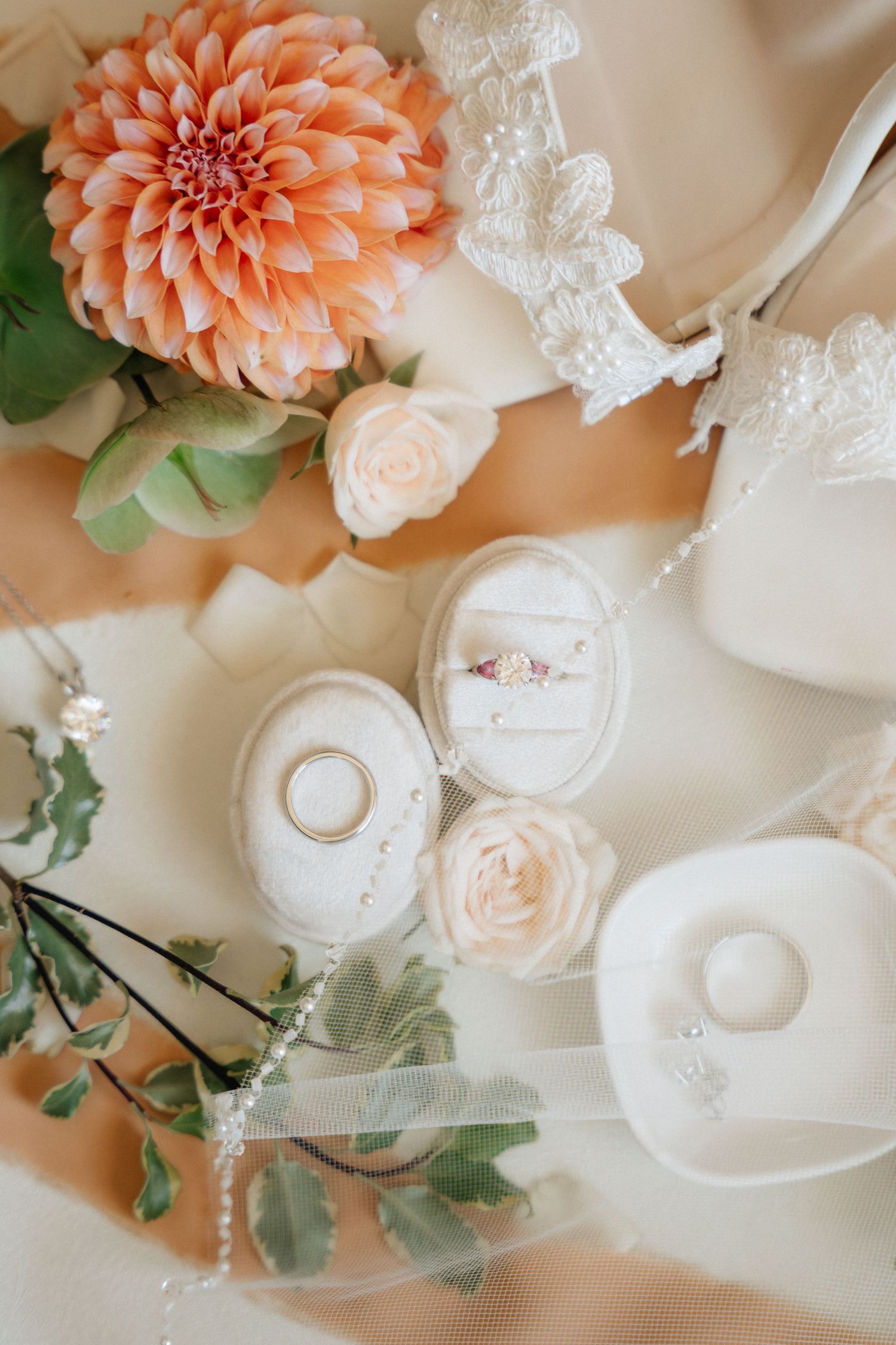 Elegant wedding flat lay with dahlia, roses, engagement ring, wedding bands, and lace details on cream fabric.