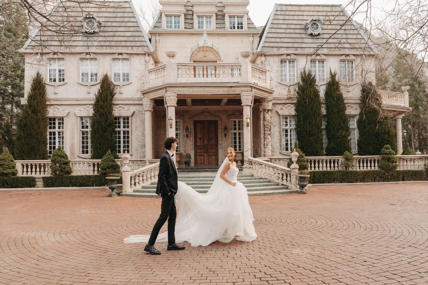 Couple in wedding attire walks along circular brick pathway before grand estate entrance.