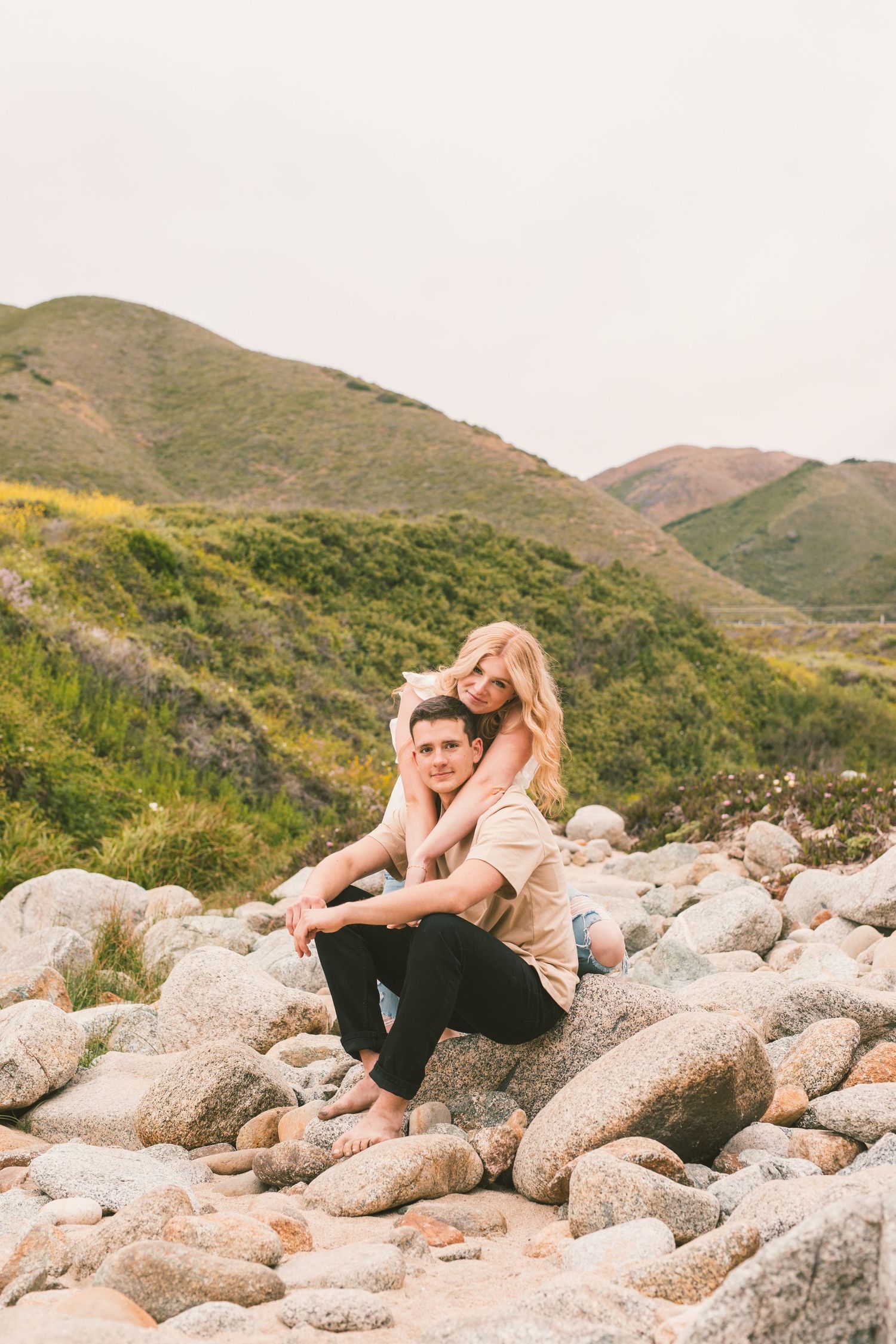 A couple shares an intimate moment while sitting on rocky terrain with rolling hills in the background.