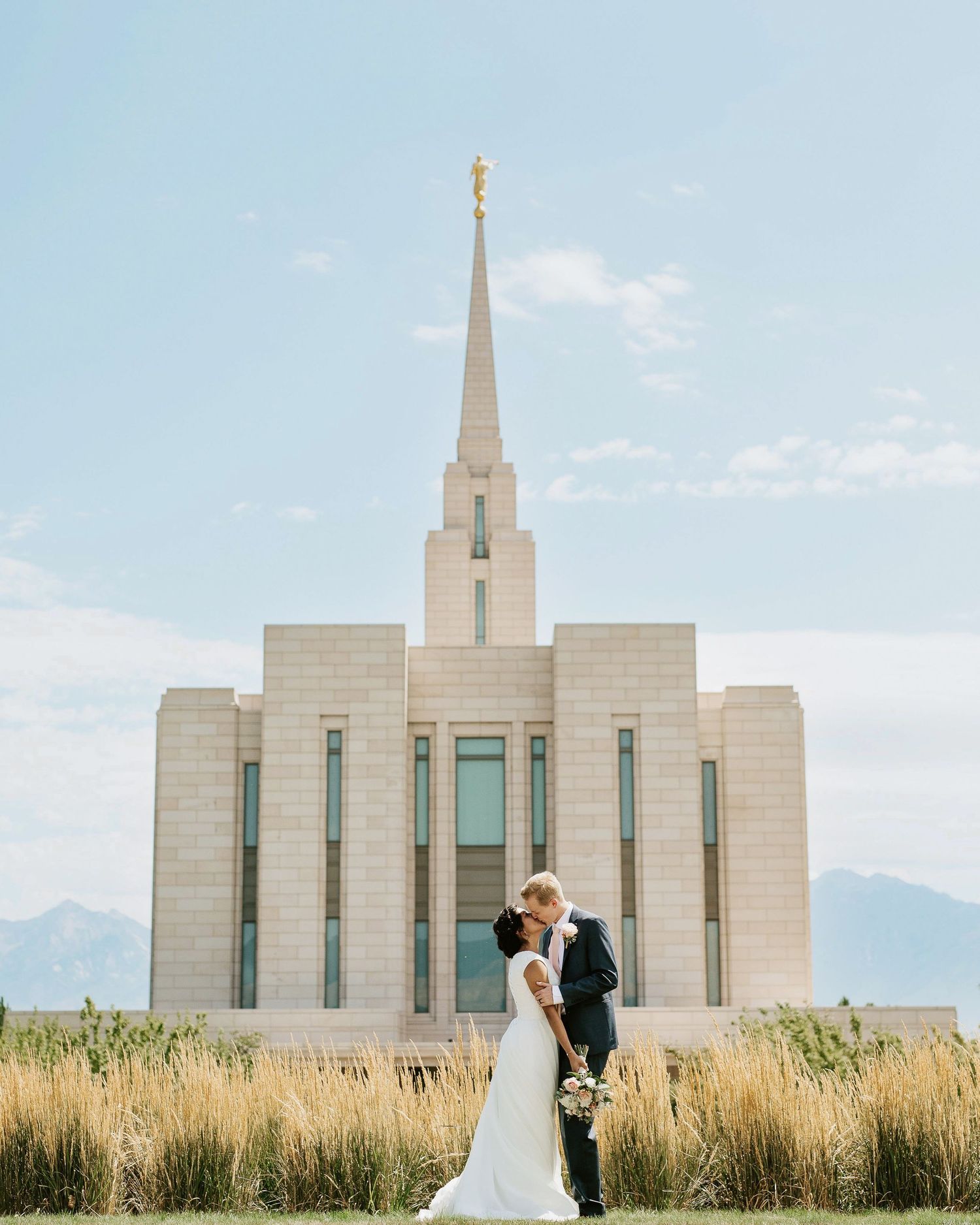 Wedding couple poses in front of a majestic white Mormon temple with mountains in the background.