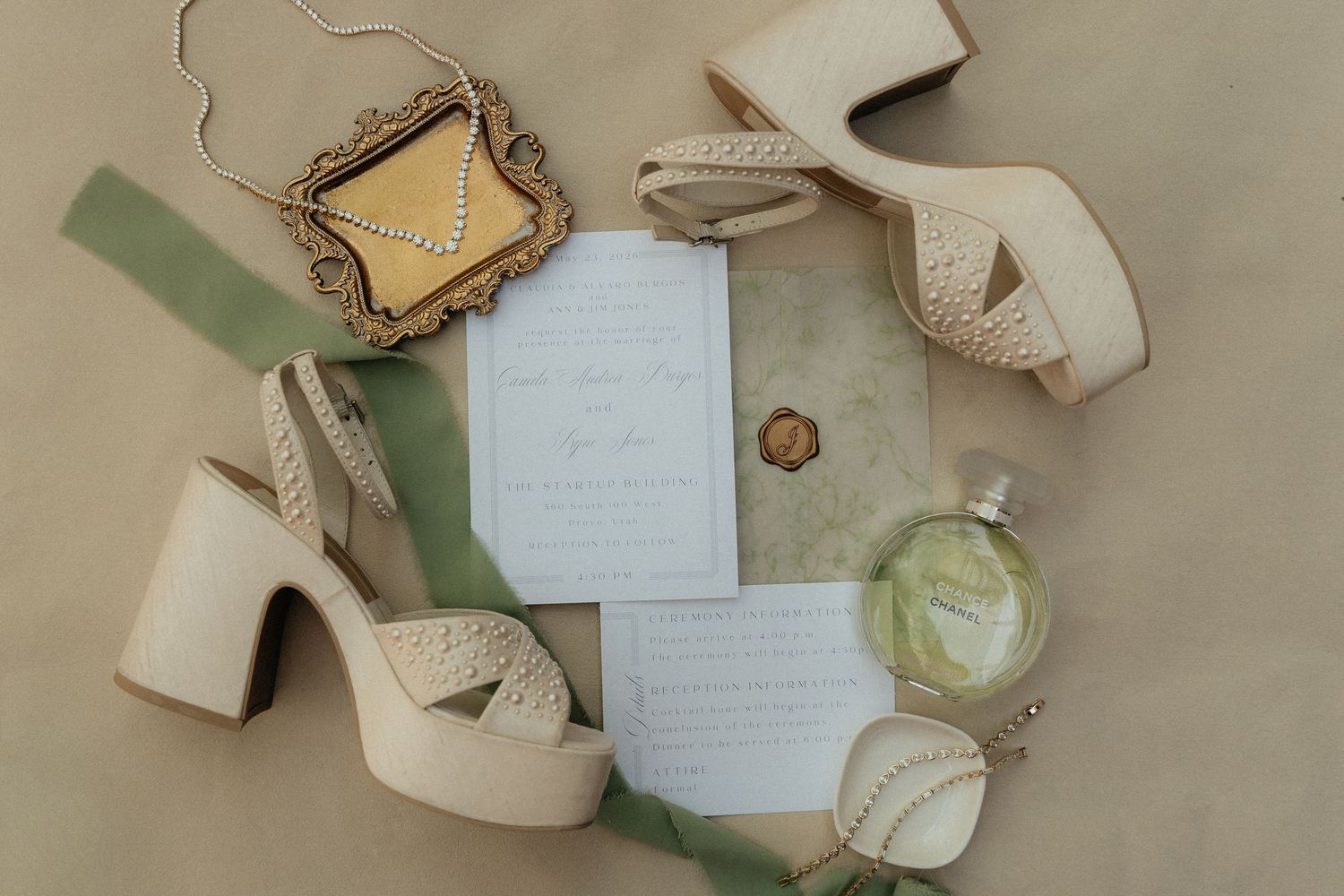 Elegant wedding flat lay with cream platform heels, floral perfume, gold jewelry box, and handwritten invitation on sage green.