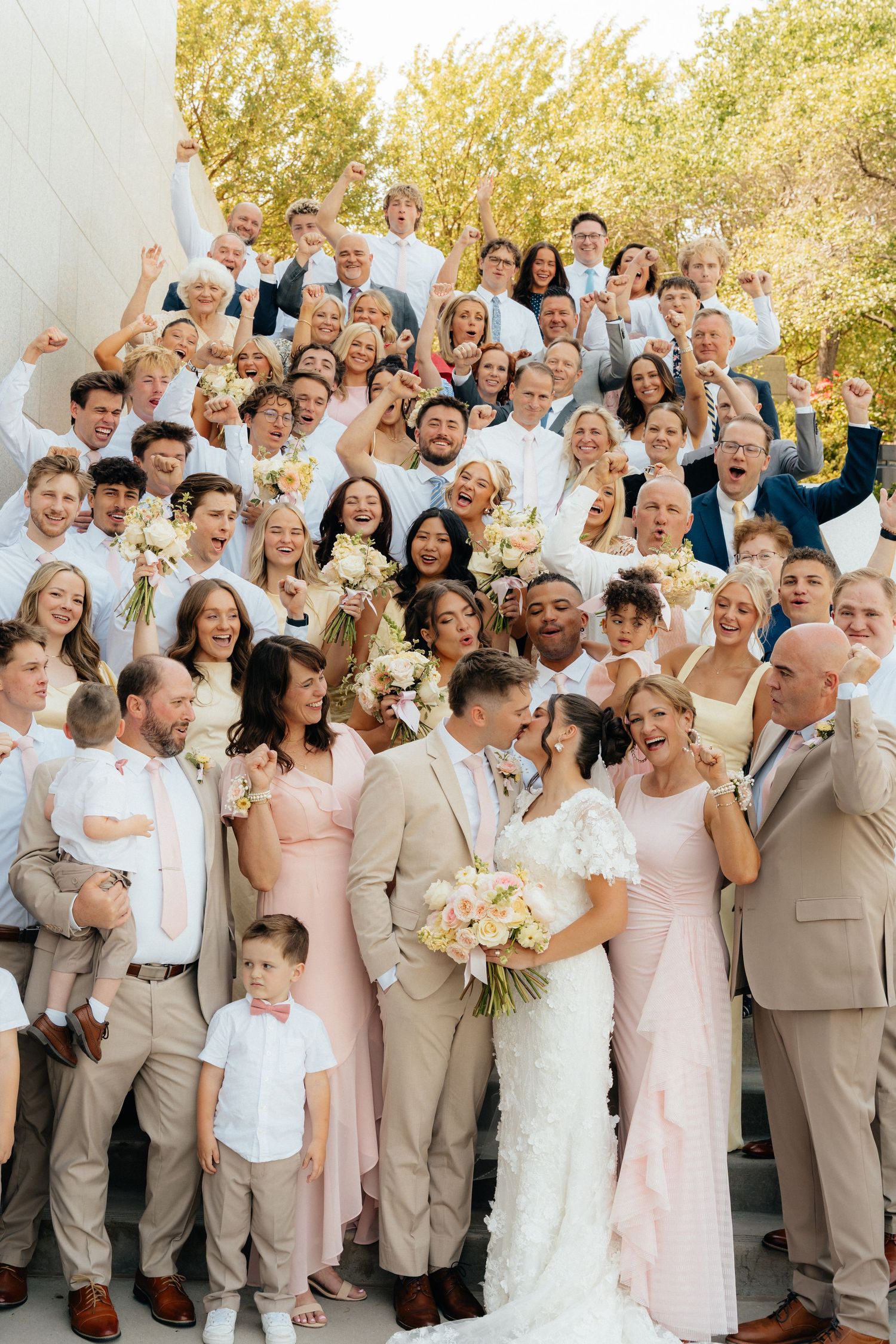 Large joyful wedding group photo on outdoor steps with bride and groom kissing, guests in blush and neutral tones.