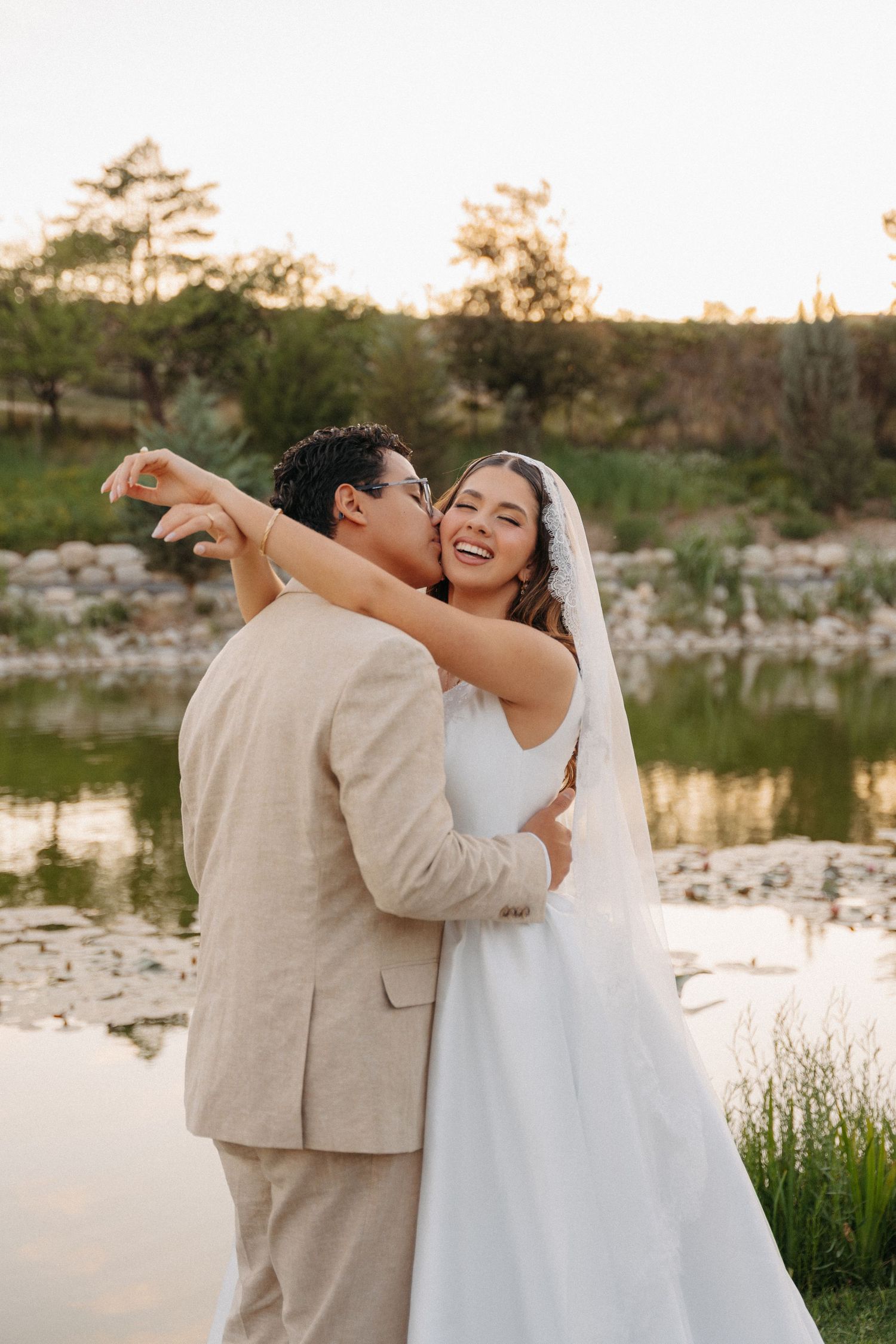 Bride and groom embracing by a pond at sunset, groom in beige suit kissing bride wearing white gown and veil.