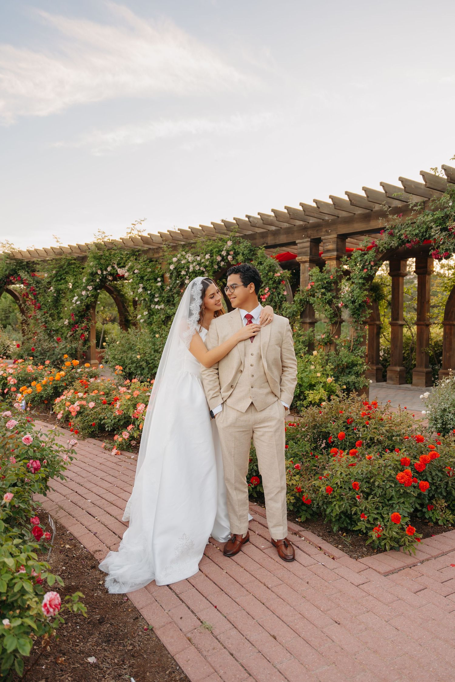 Bride and groom pose in a rose garden pergola, bride in white gown with veil, groom in tan suit.