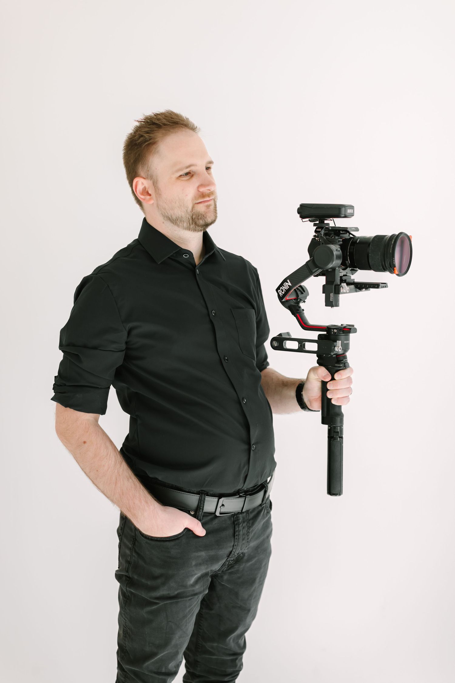 A man with a beard holding a professional video camera on a stabilizer in front of a plain white background.