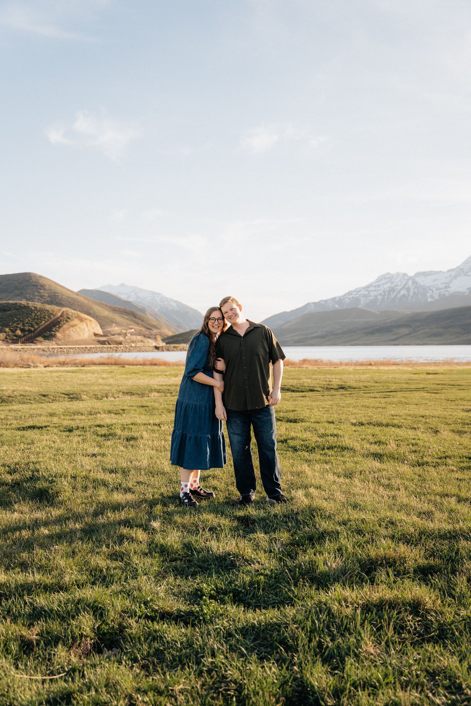 Couple smiling and embracing on green grass field with snow-capped mountains and lake in background on sunny day.