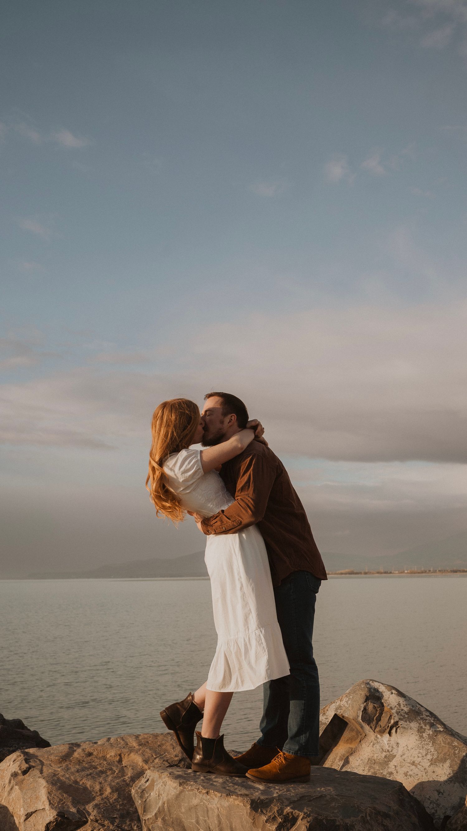 Romantic couple embracing on rocky shoreline during golden hour photoshoot.