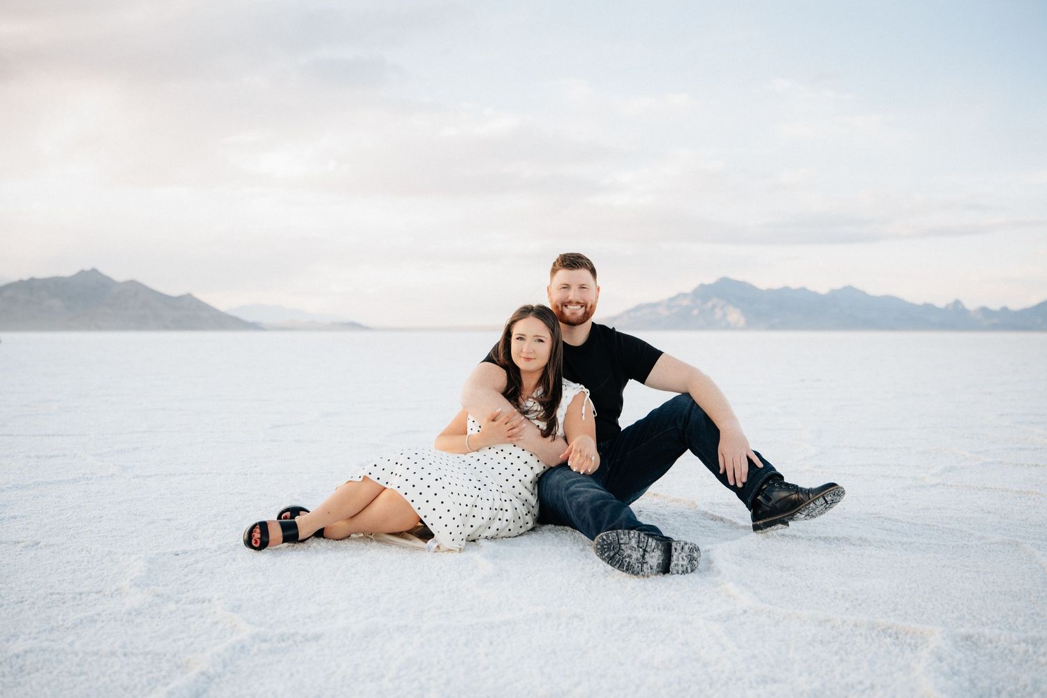 Couple sitting together on white salt flats with mountain backdrop during engagement photo session.