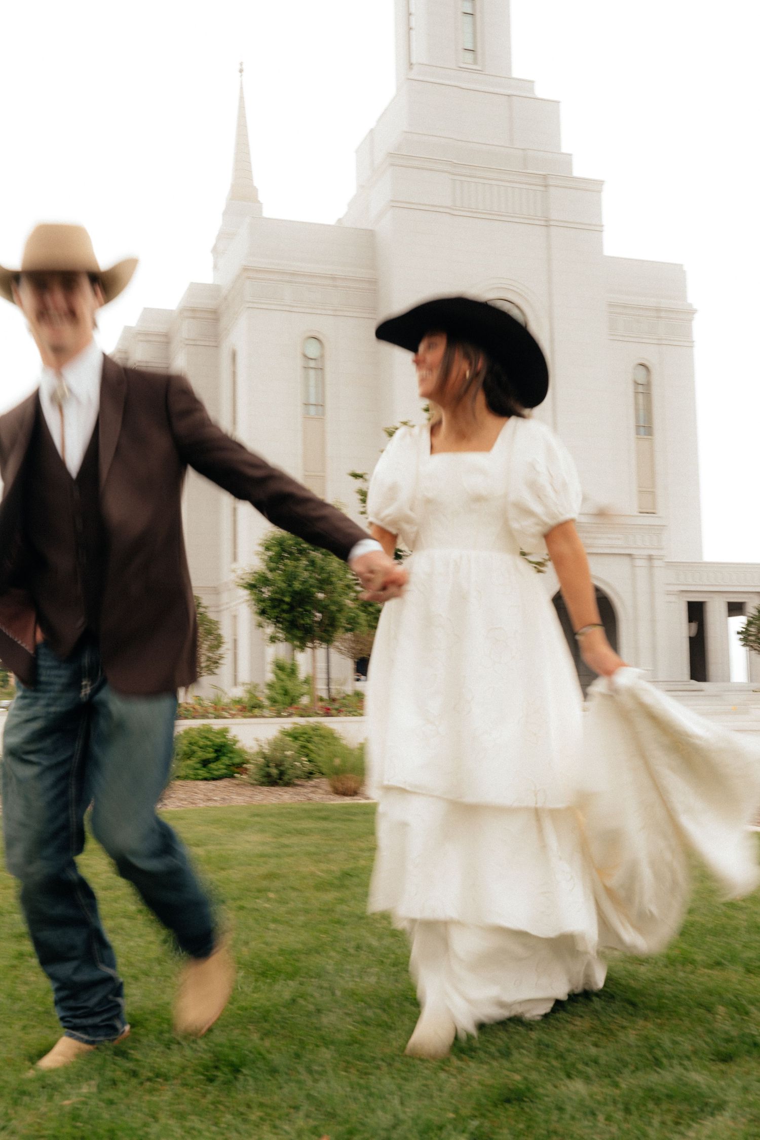 Bride in white dress and cowboy hat holds groom's hand outside a white LDS temple on their wedding day.