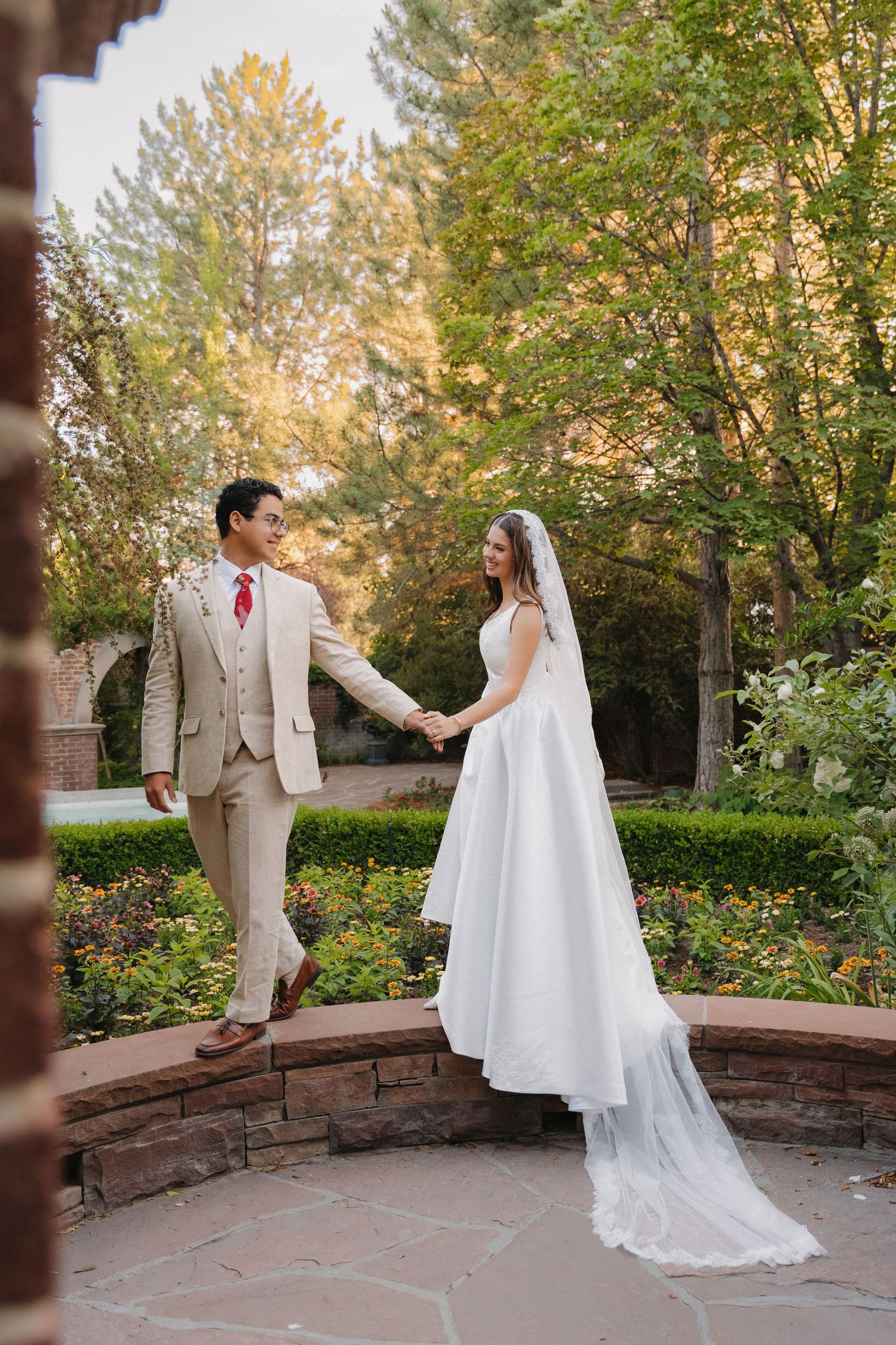 Bride and groom in beige suit hold hands in a lush garden, bride's cathedral veil trailing on stone patio.