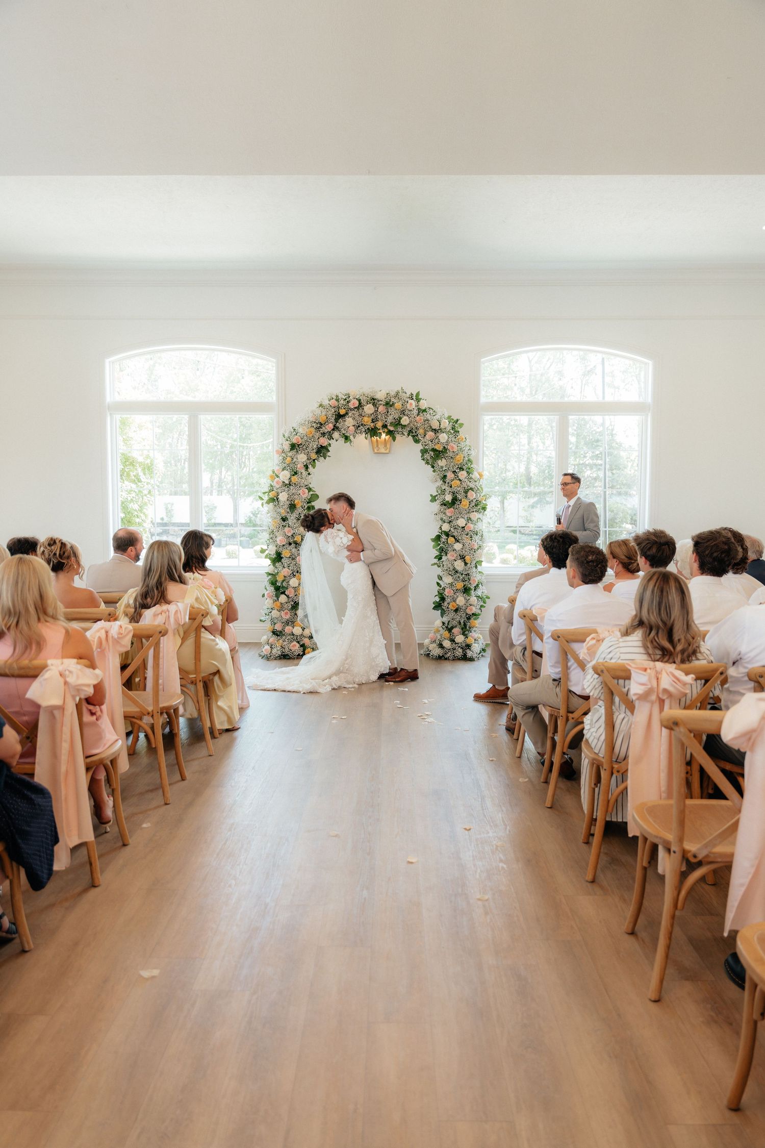 Bride and groom share first kiss at elegant indoor ceremony beneath a lush floral arch with seated guests.