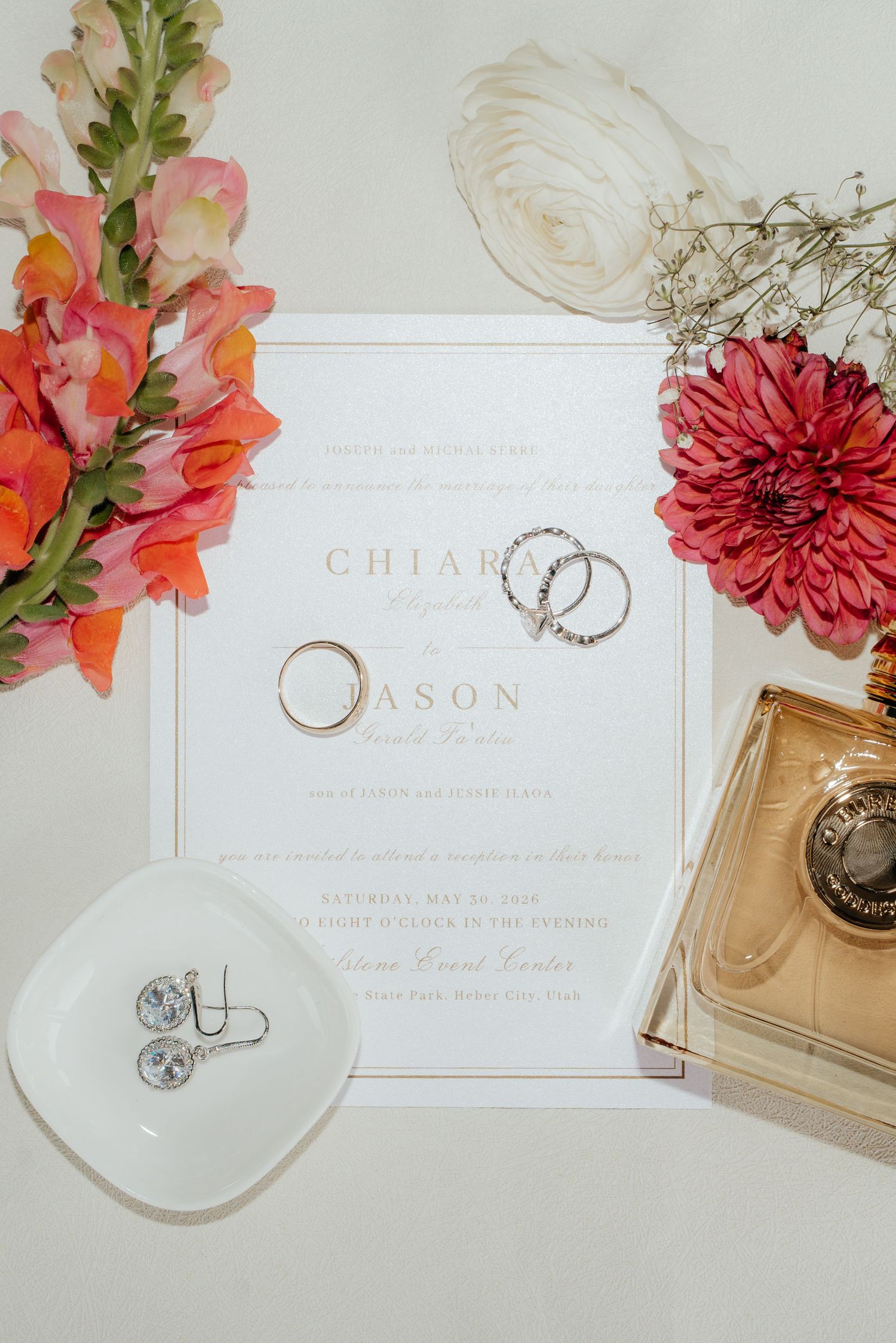 Wedding flat lay with invitation for Chiara and Jason, gold rings, diamond earrings, perfume bottle, and pink florals.