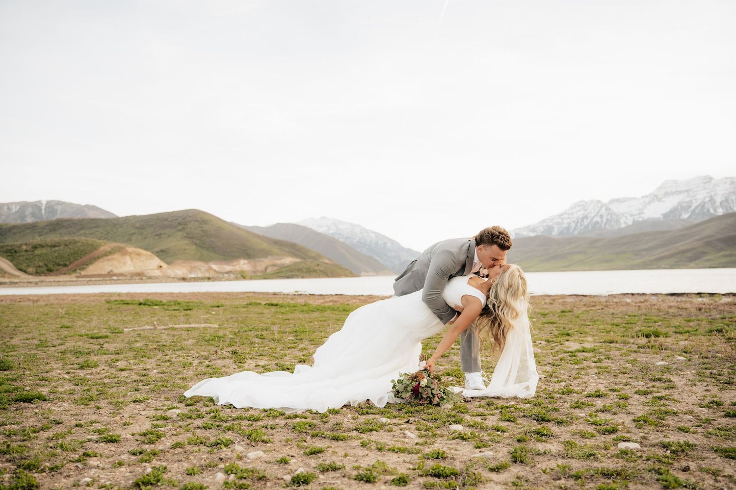 Bride and groom share a romantic dip kiss outdoors with snow-capped mountains and a lake in the background.