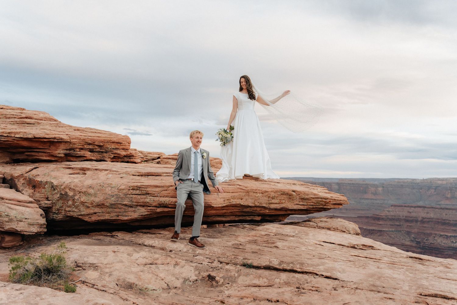 Bride and groom pose on red rock canyon cliffs during a stunning desert elopement ceremony at sunset.