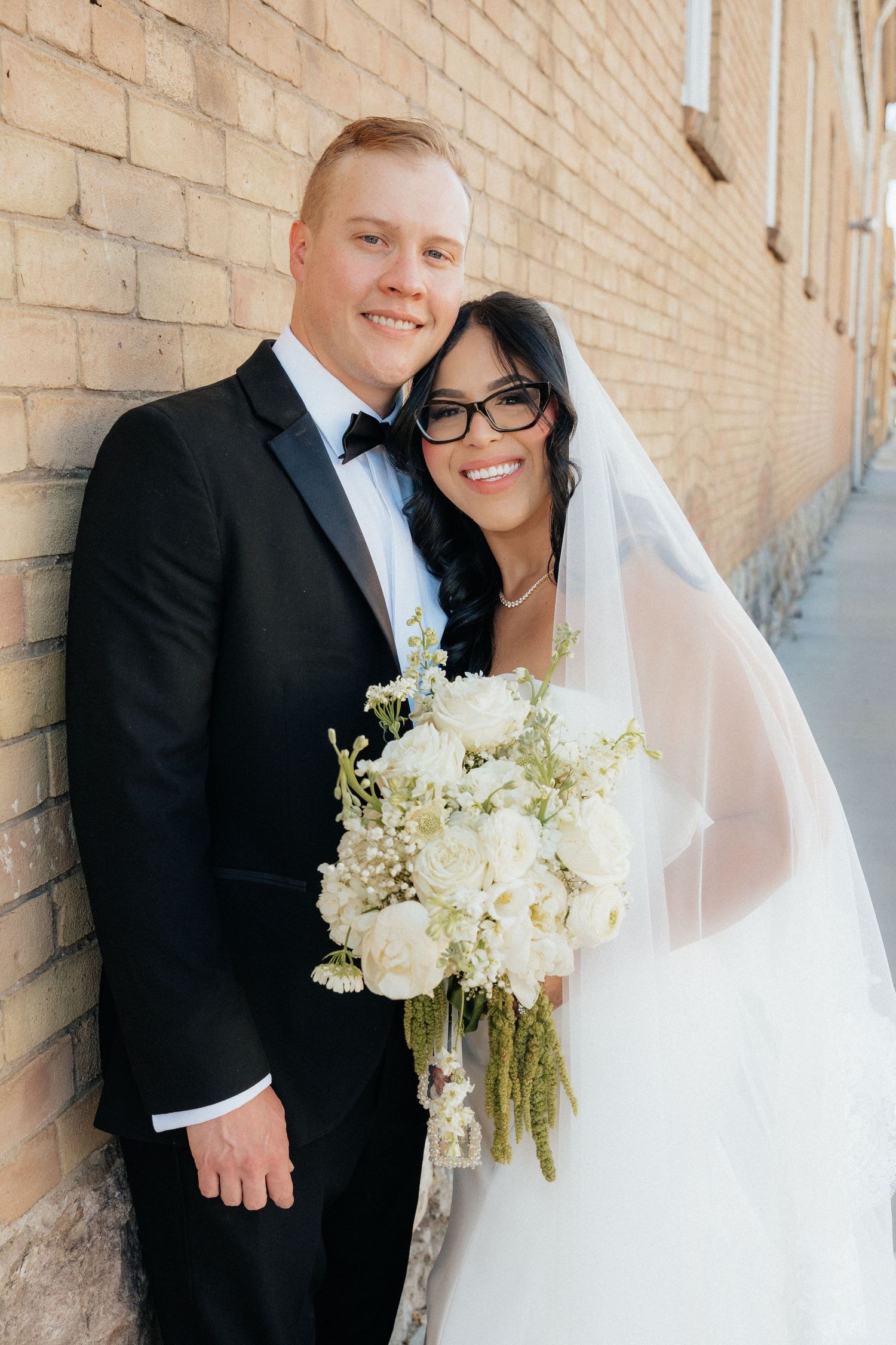 Bride in white gown and veil holding white bouquet poses with groom in black tuxedo against brick wall.