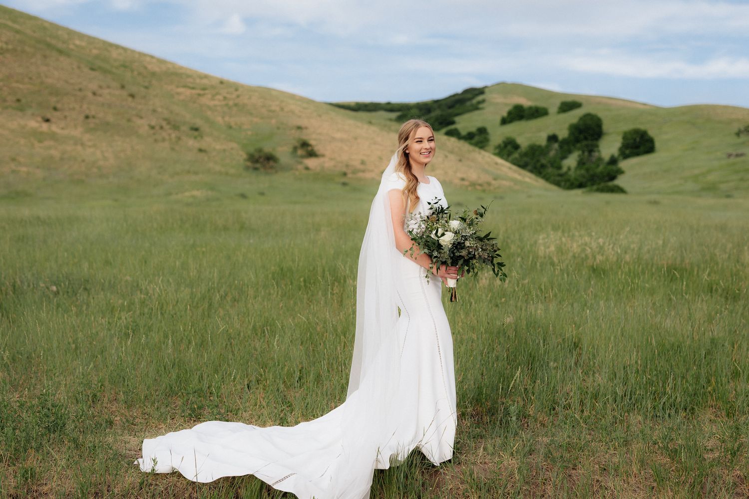 A bride in a flowing white dress holds a wildflower bouquet in a green meadow.