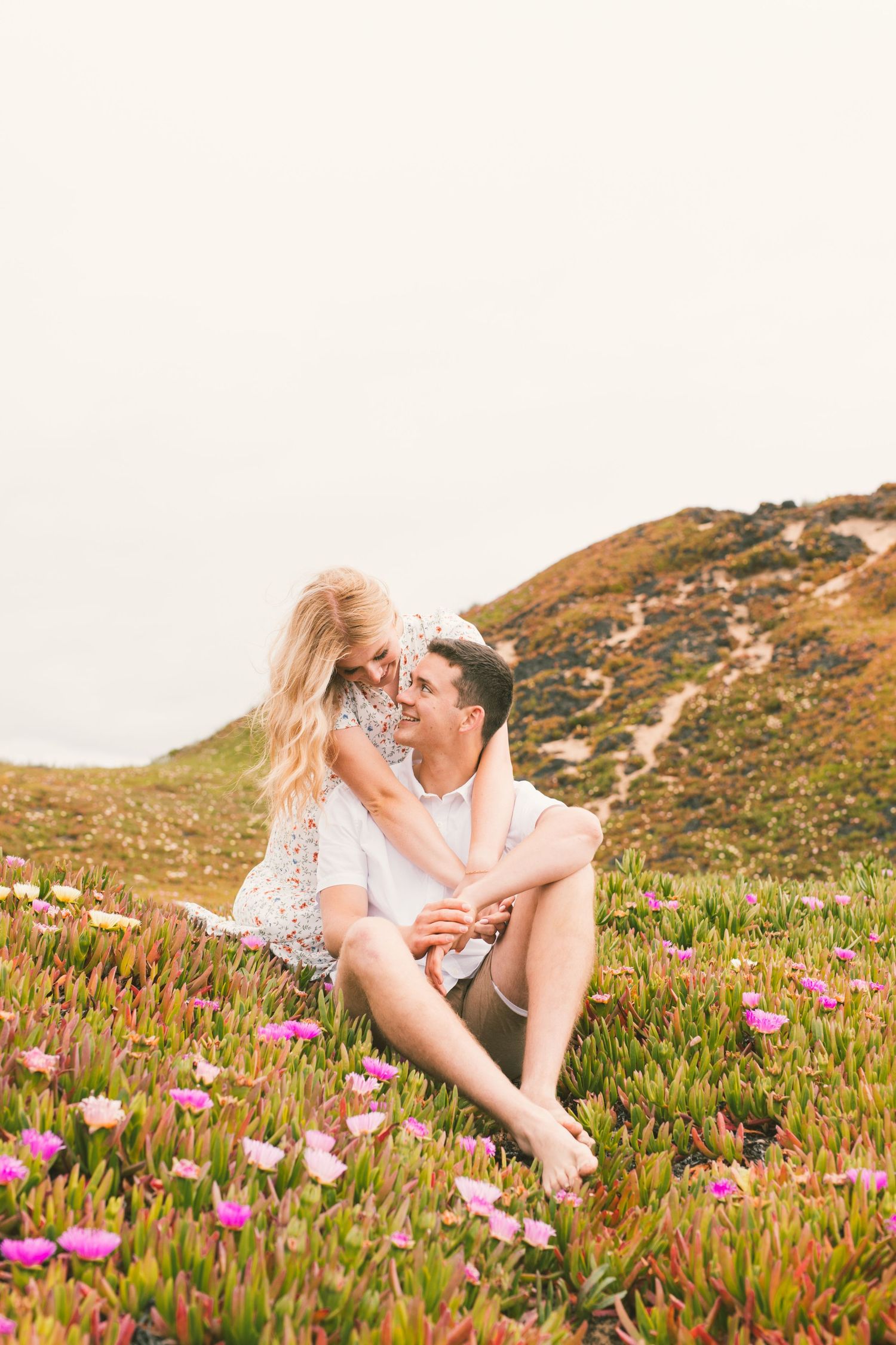 Couple lounging intimately in field of pink coastal flowers during golden hour.