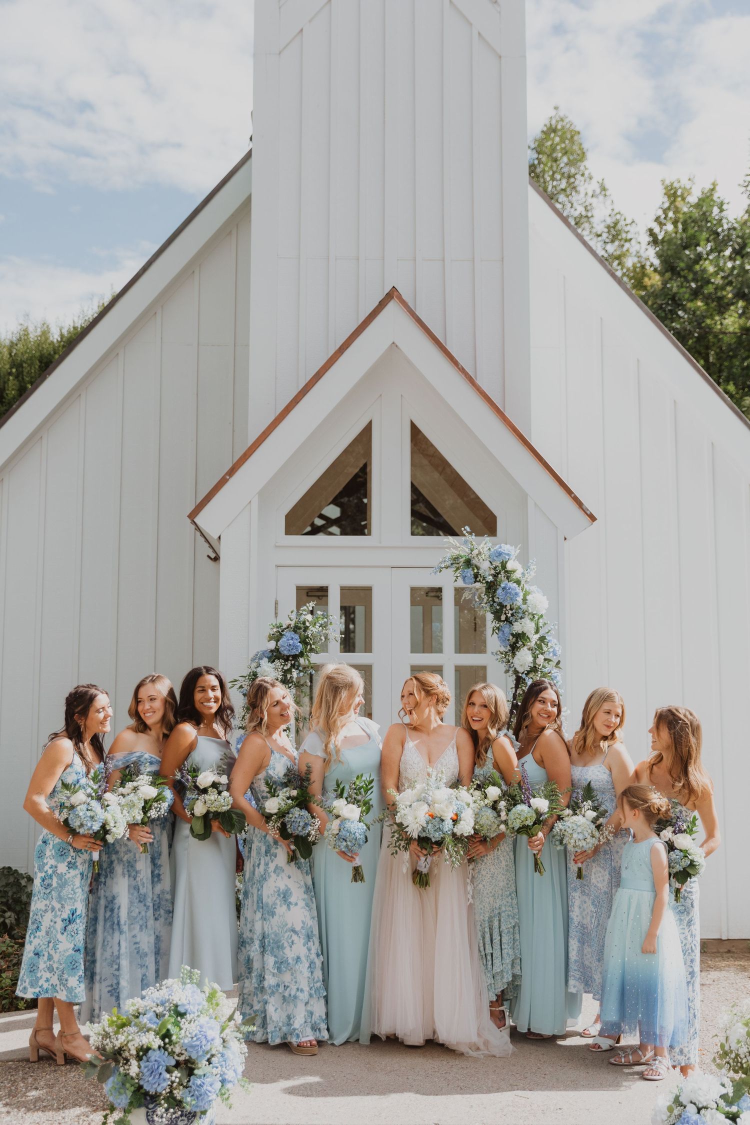 Bridesmaids in varying shades of blue dresses pose in front of white chapel.