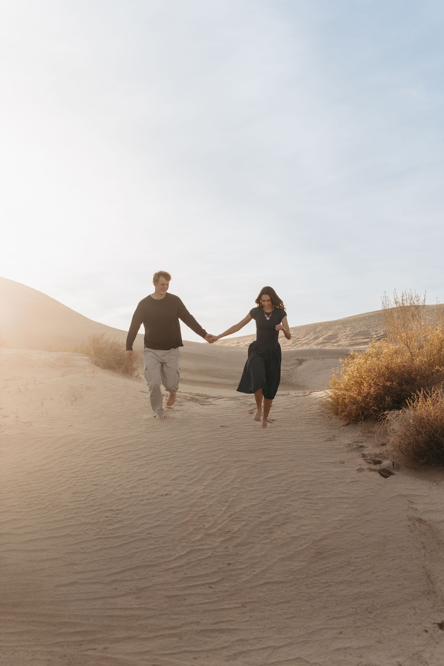 A couple holding hands while walking down a sandy dune slope at sunset with golden light and desert brush nearby.