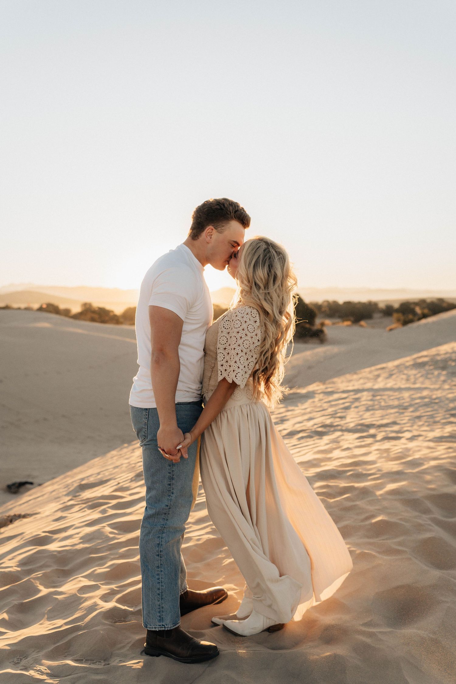 Two people share an intimate moment while kneeling in the sand during a warm sunset.