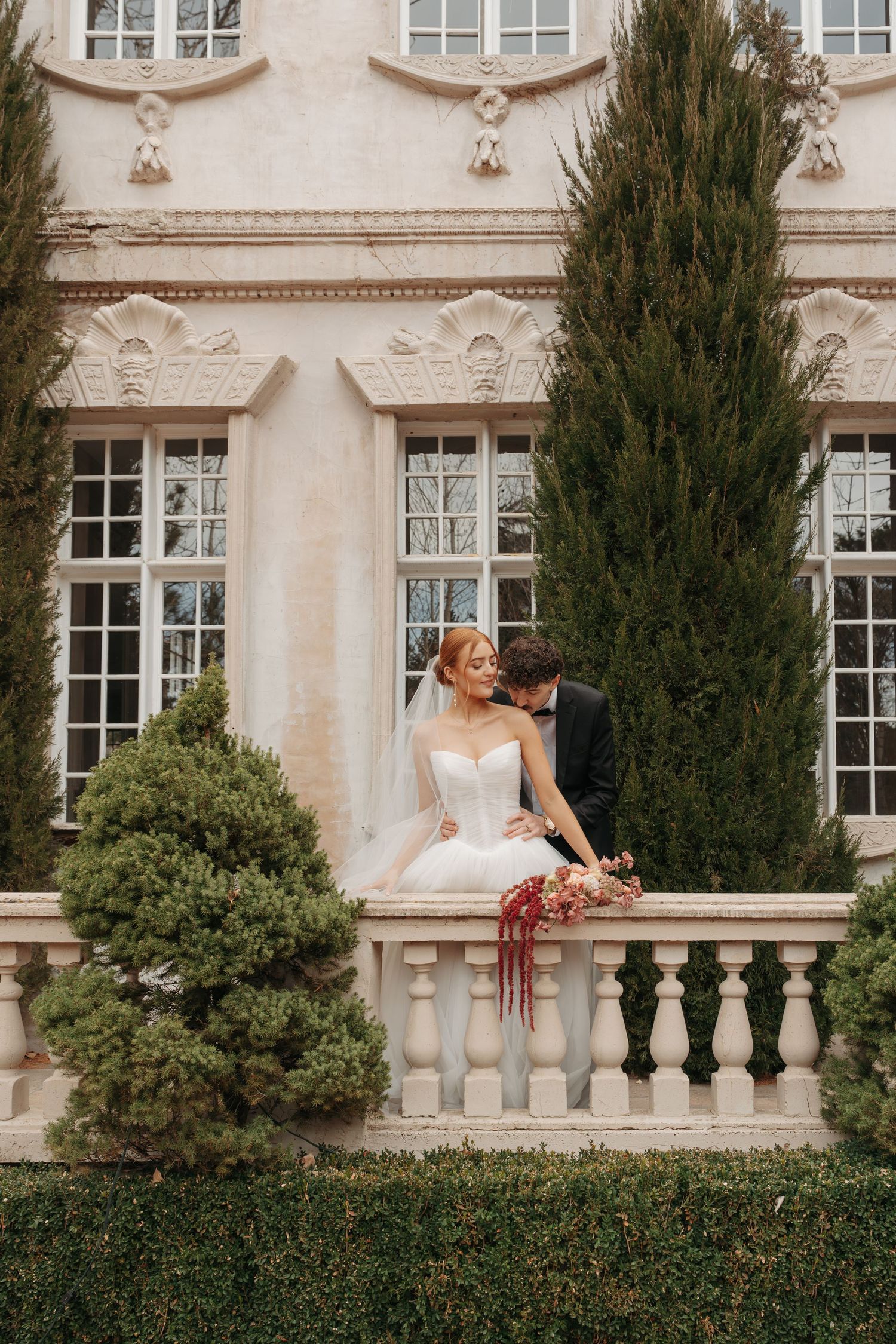 Wedding couple embraces on an elegant stone balcony with cypress trees and classic architecture.