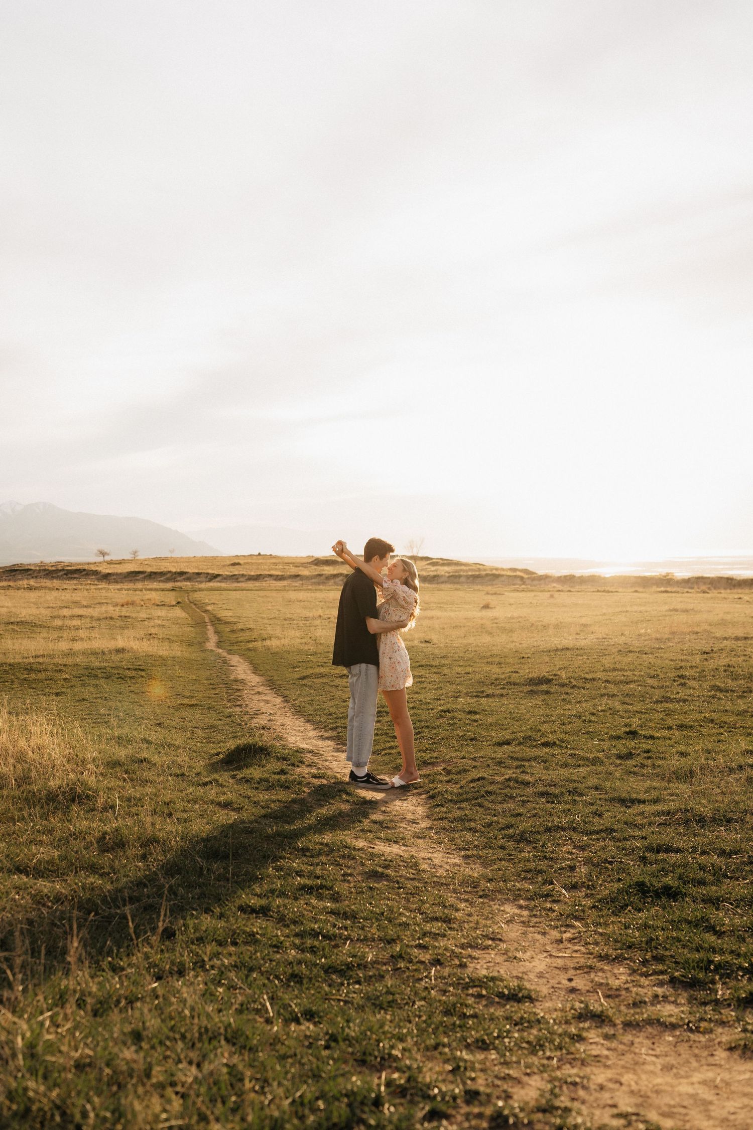 Couple embracing and kissing in a golden sunlit field with a winding dirt path and mountains in the background.
