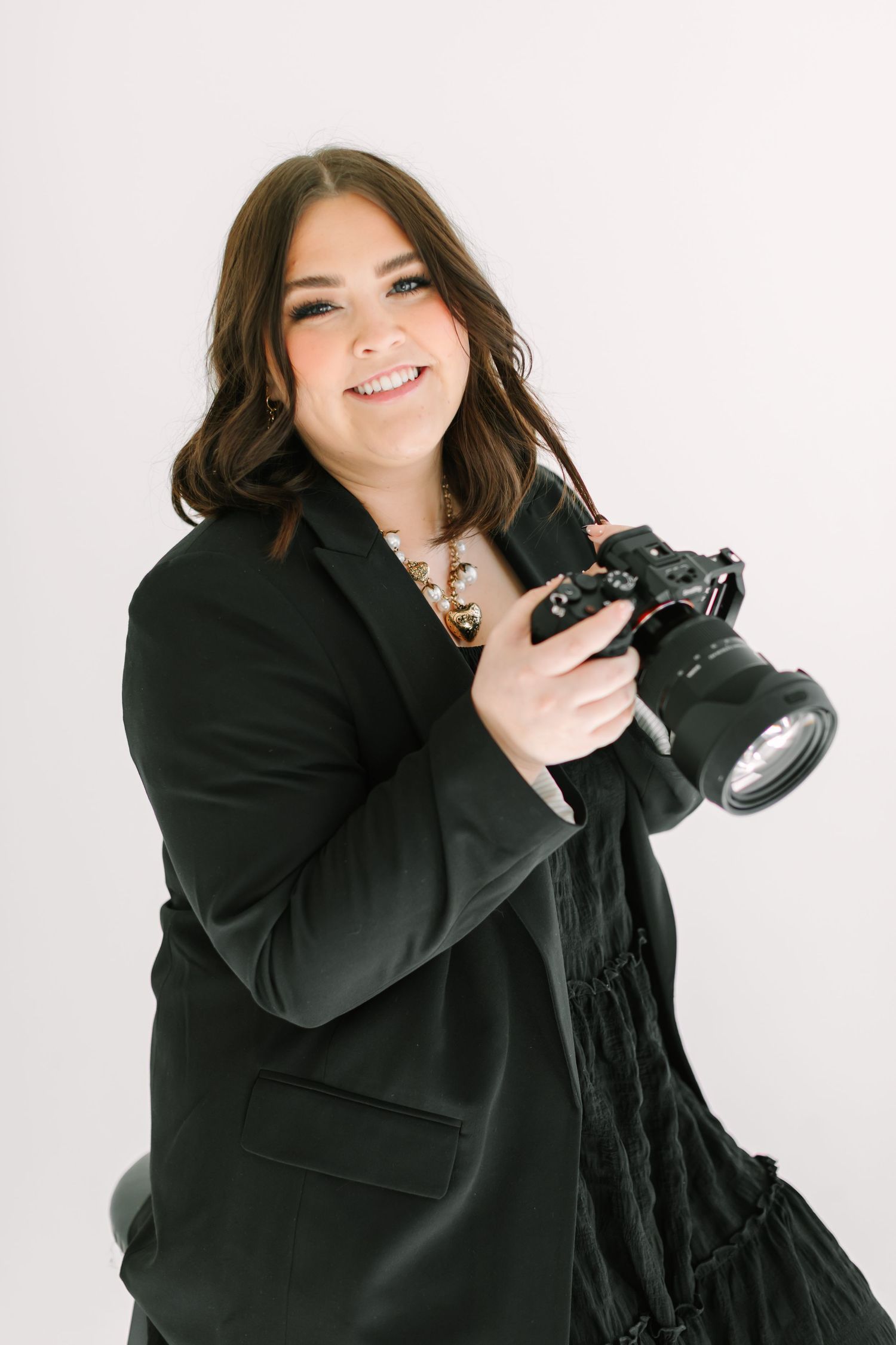 A smiling woman wearing a black coat and holding a camera stands against a white background.