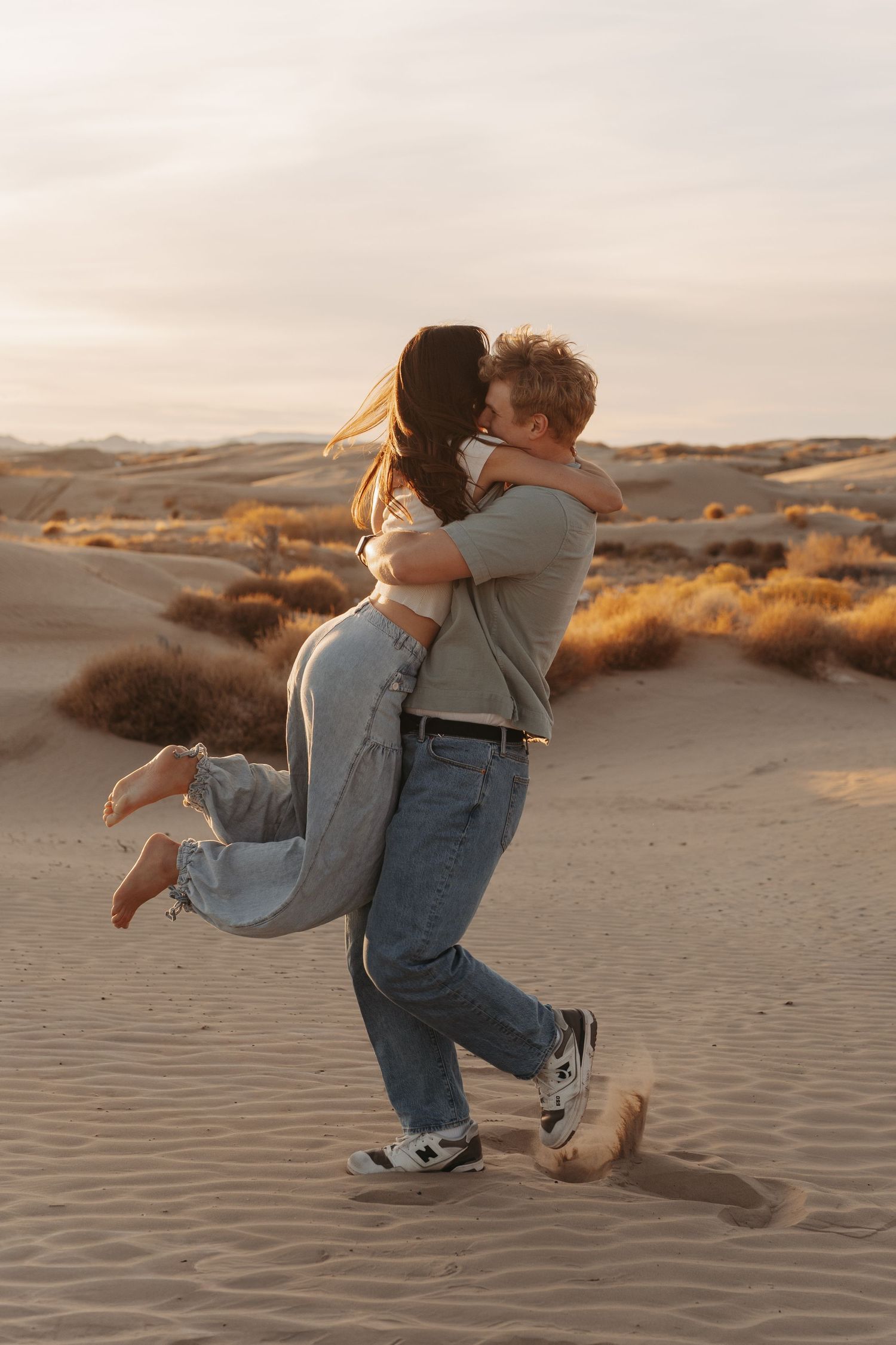 Romantic couple embracing and spinning in sand dunes at sunset with warm golden light.