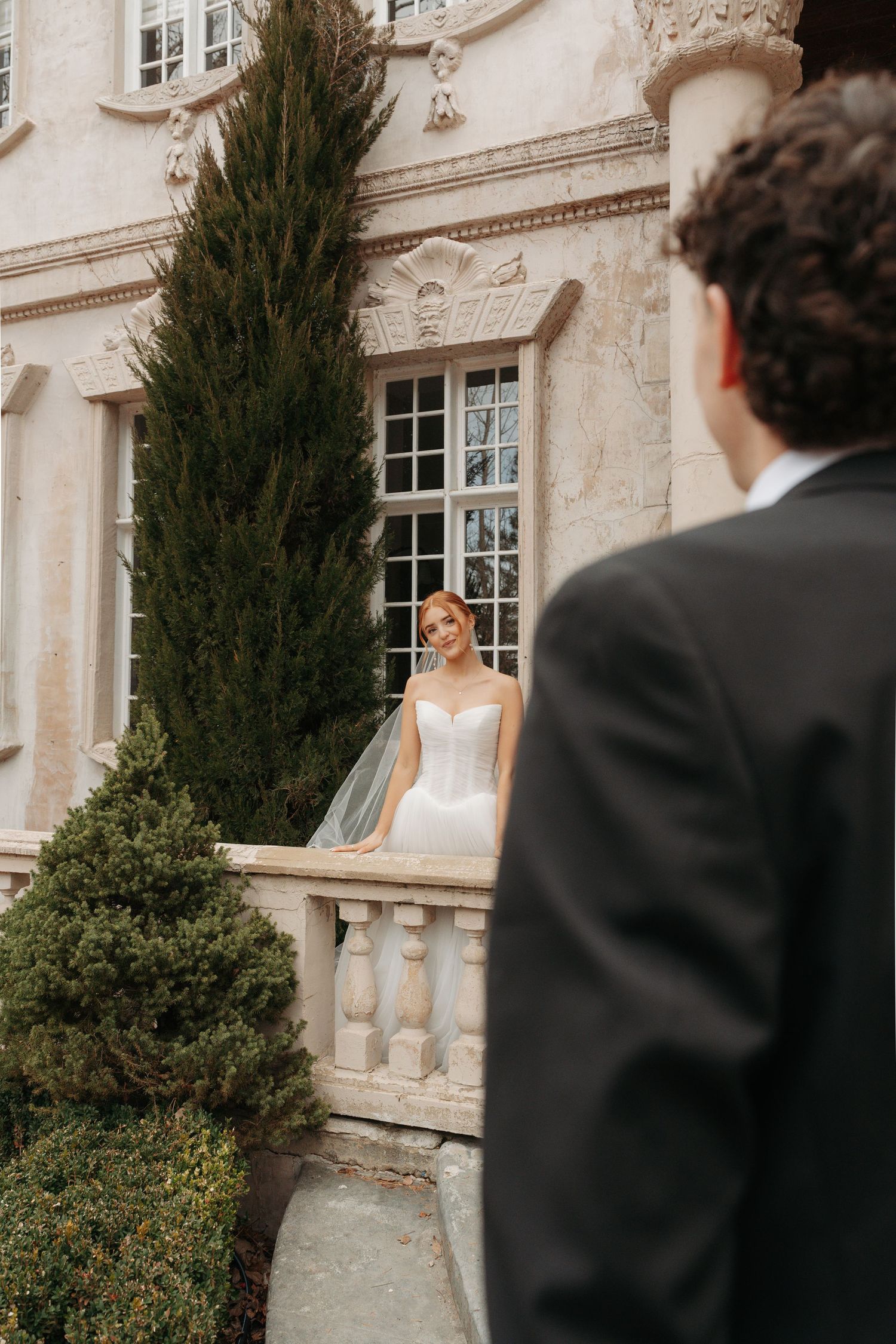 A romantic wedding scene with a couple in formal attire standing near cypress trees and a grand stone building.