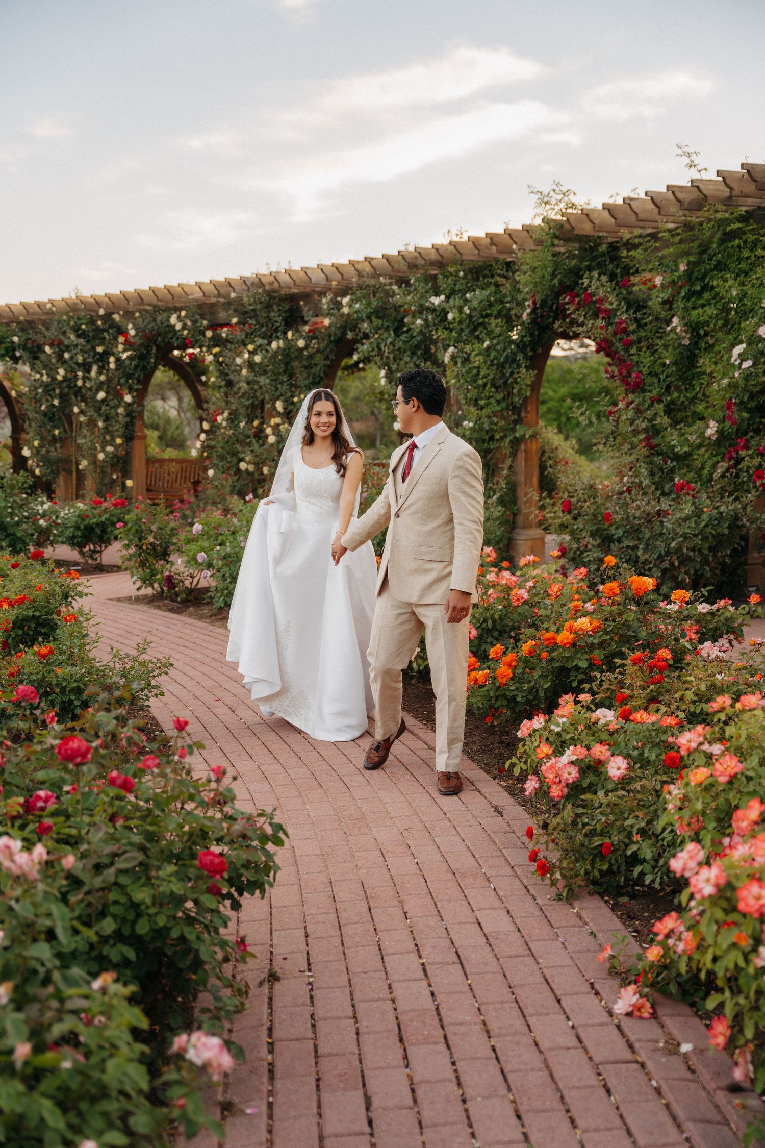 Bride and groom walk hand-in-hand through a vibrant rose garden pergola on their wedding day.