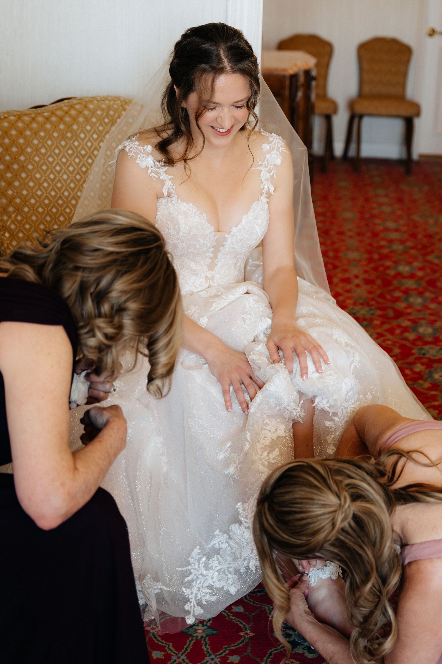 Smiling bride in lace wedding gown being helped by bridesmaids with garter on red carpet floor.