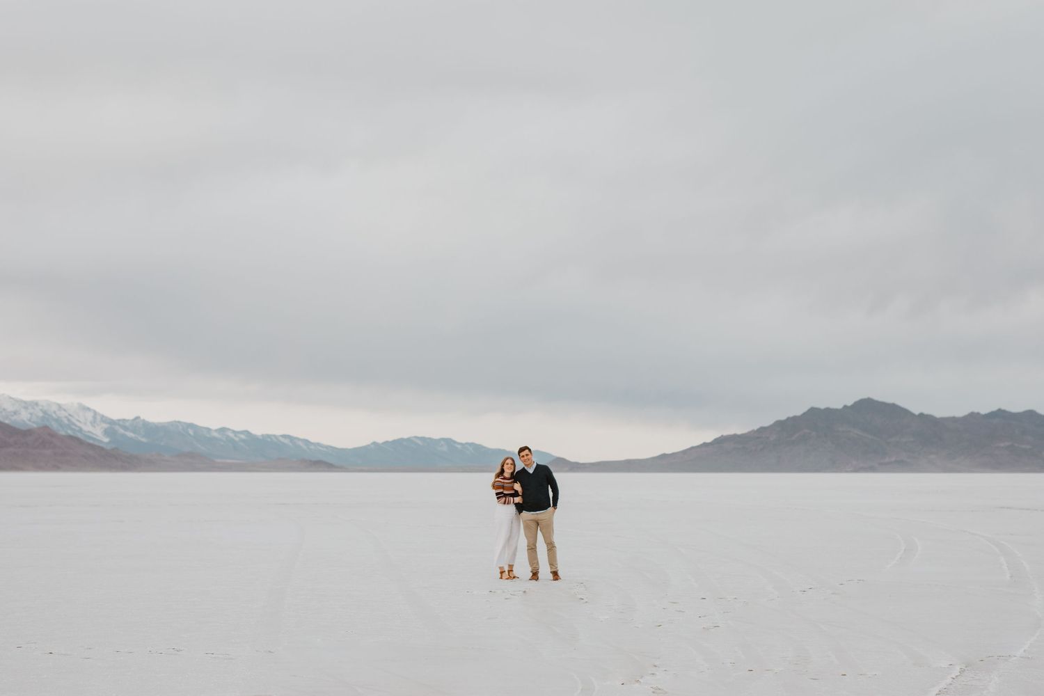 Couple embracing on scenic salt flats with mountains in background under cloudy sky.