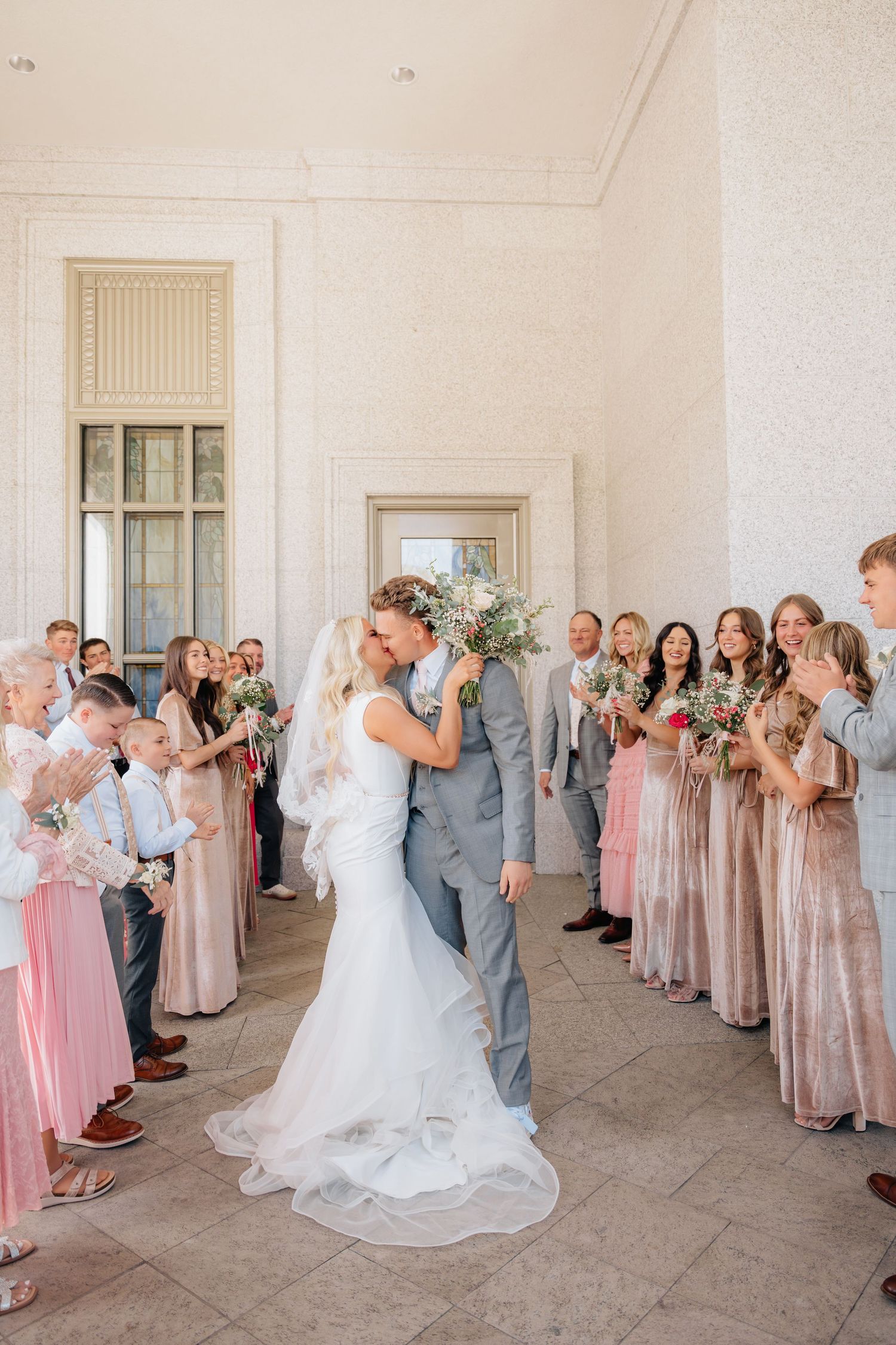 Bride and groom share a kiss surrounded by wedding party in pink dresses and gray suits in elegant venue.