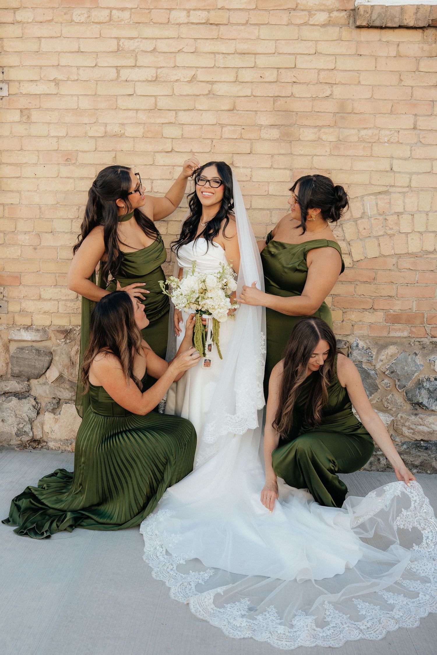 Bride in white gown holds bouquet surrounded by four bridesmaids in olive green dresses against a brick wall.