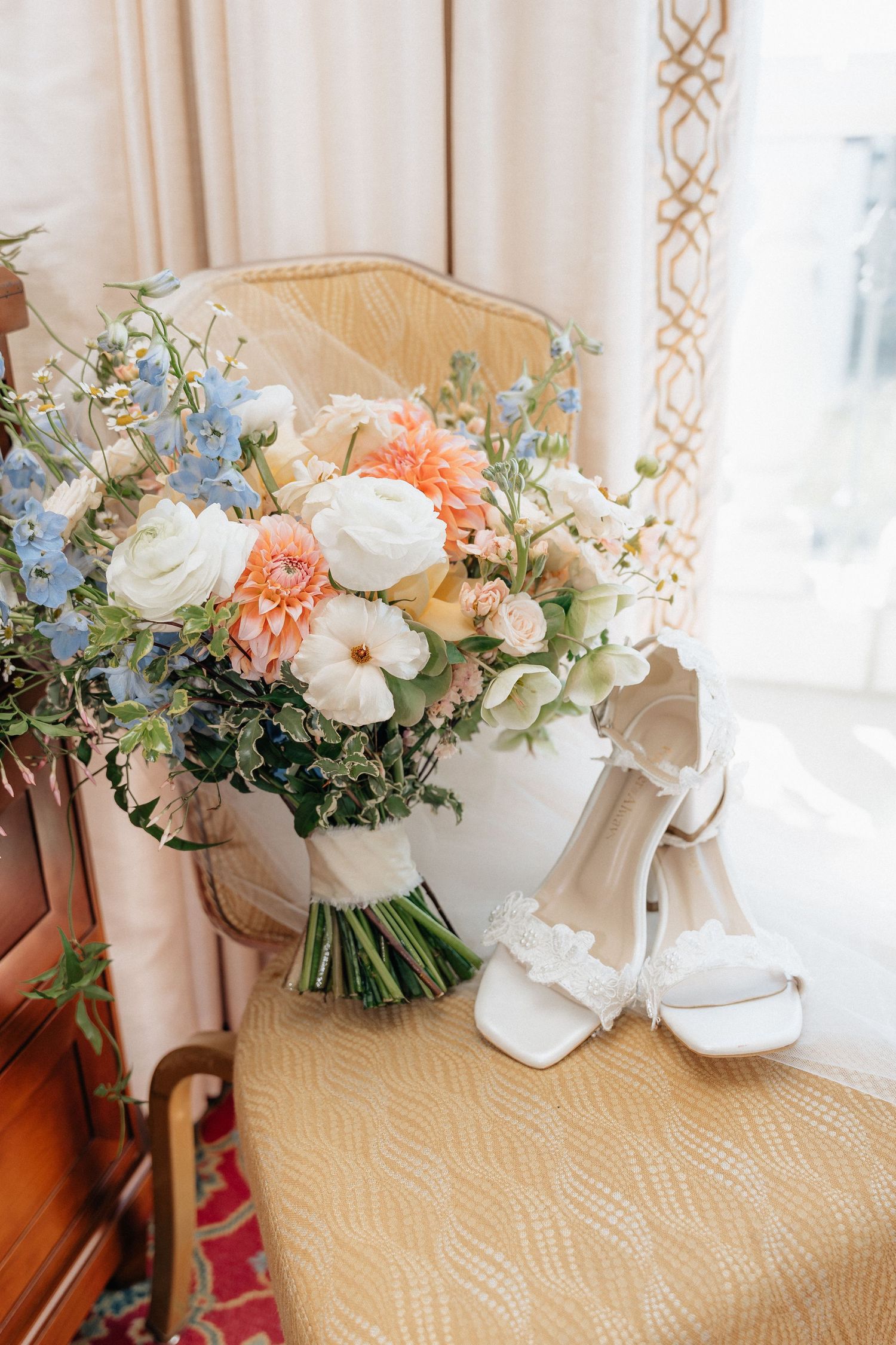 Bridal bouquet with peach, white, and blue flowers beside white heeled wedding shoes on a gold chair.