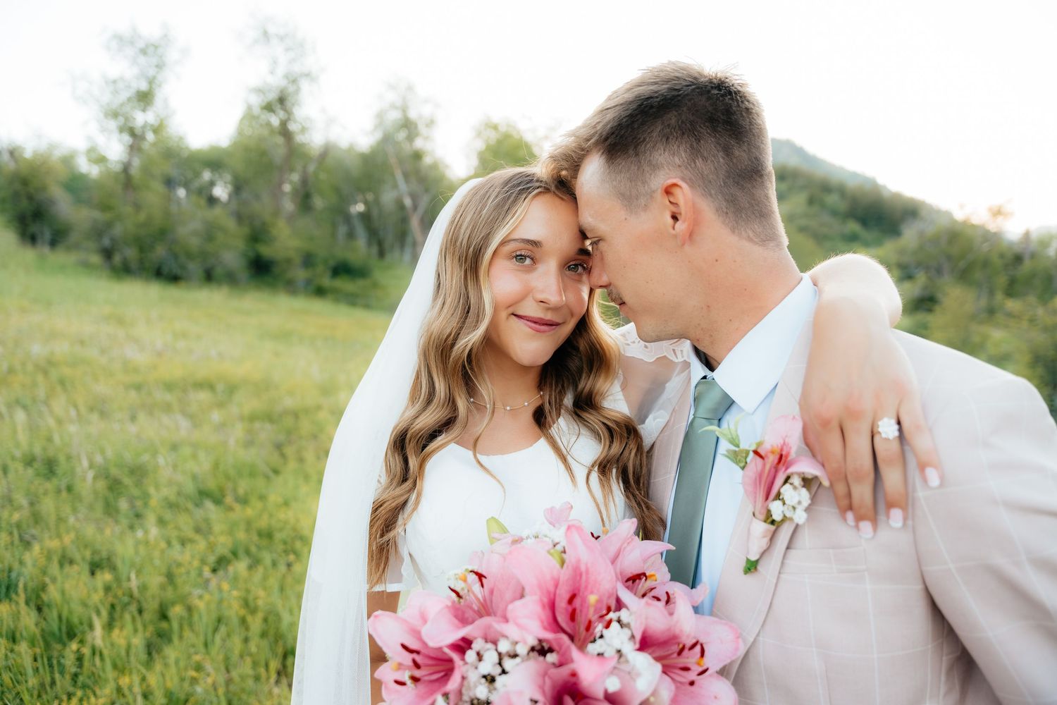 Bride and groom in a green meadow, sharing a tender moment, bride holding pink lily bouquet in wedding attire.