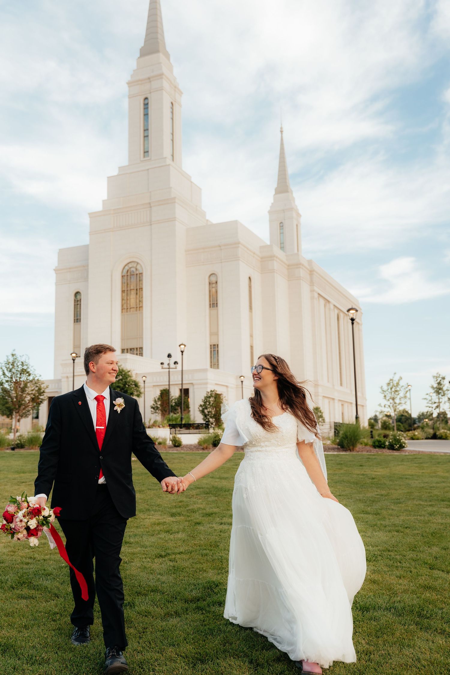 Bride and groom holding hands walking on green grass in front of a white LDS temple on their wedding day.