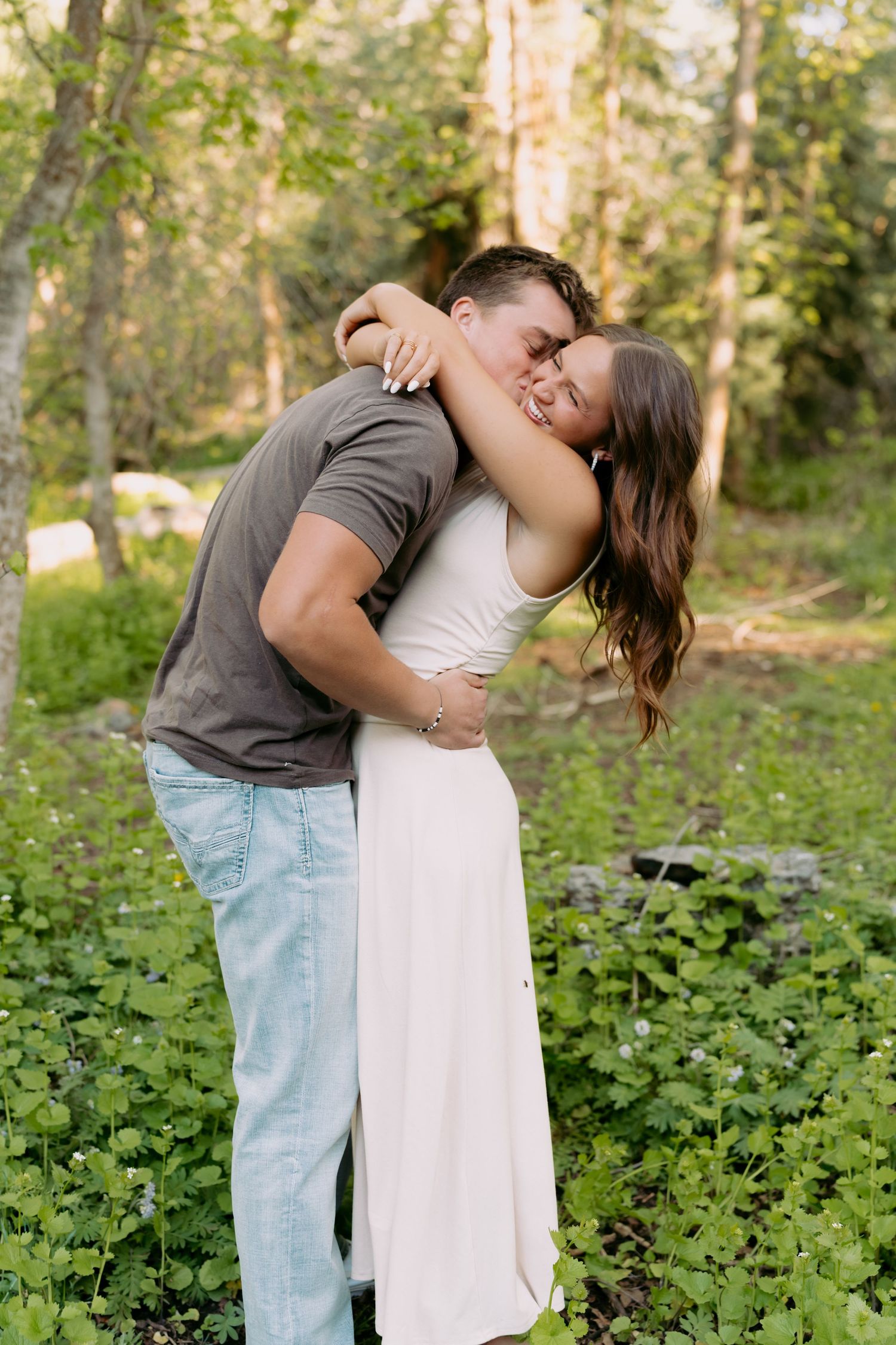 A couple embraces and kisses in a lush green forest setting, the woman in a white dress hugging the man in a gray shirt.