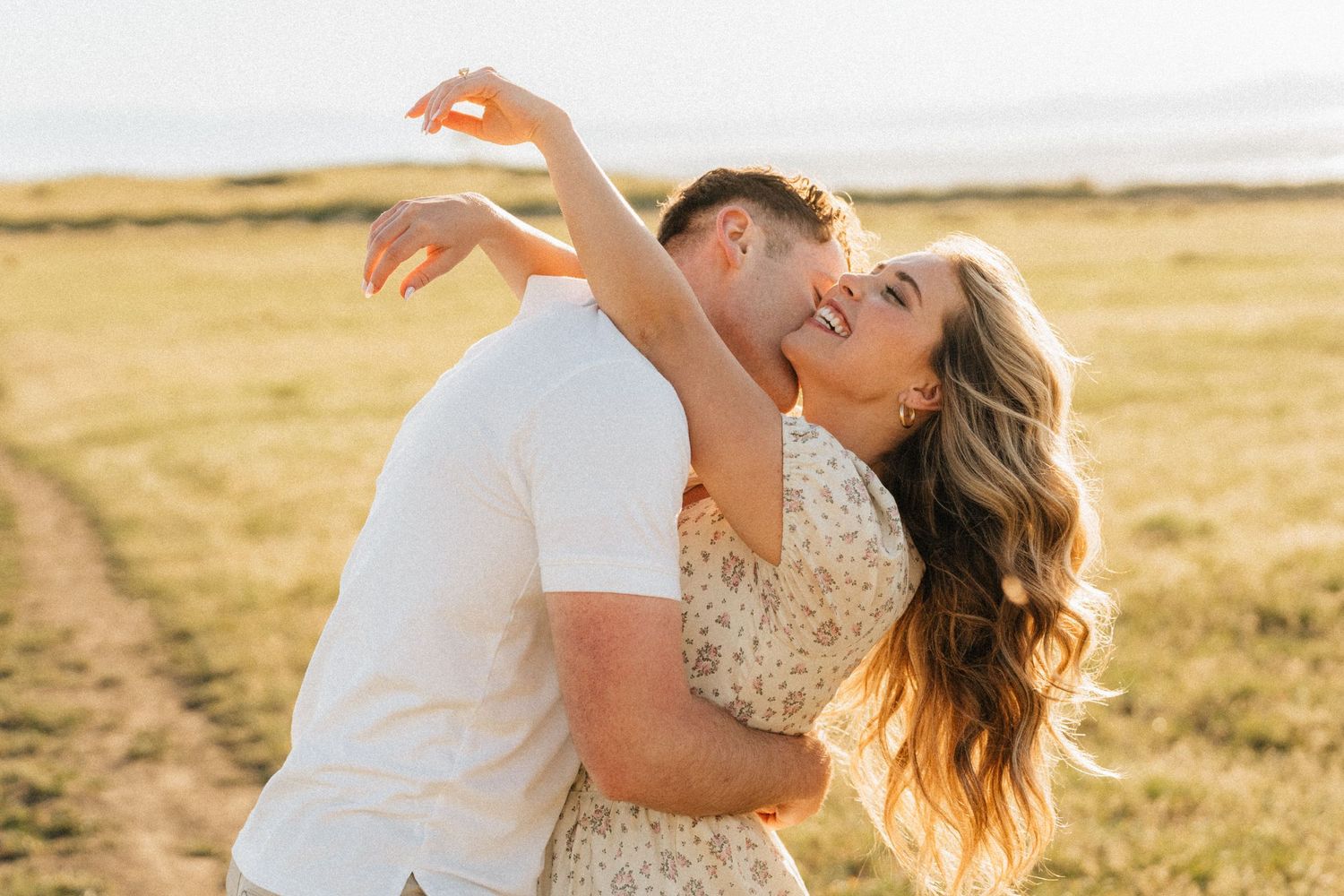 Couple sharing a romantic dance in a sunny golden field, woman in floral dress with arm raised, man in white shirt.