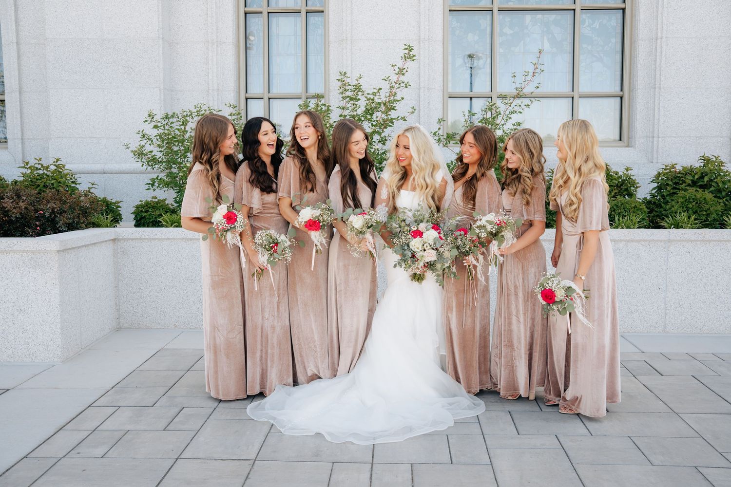 Bride in white gown poses with bridesmaids in champagne dresses holding colorful bouquets outdoors.