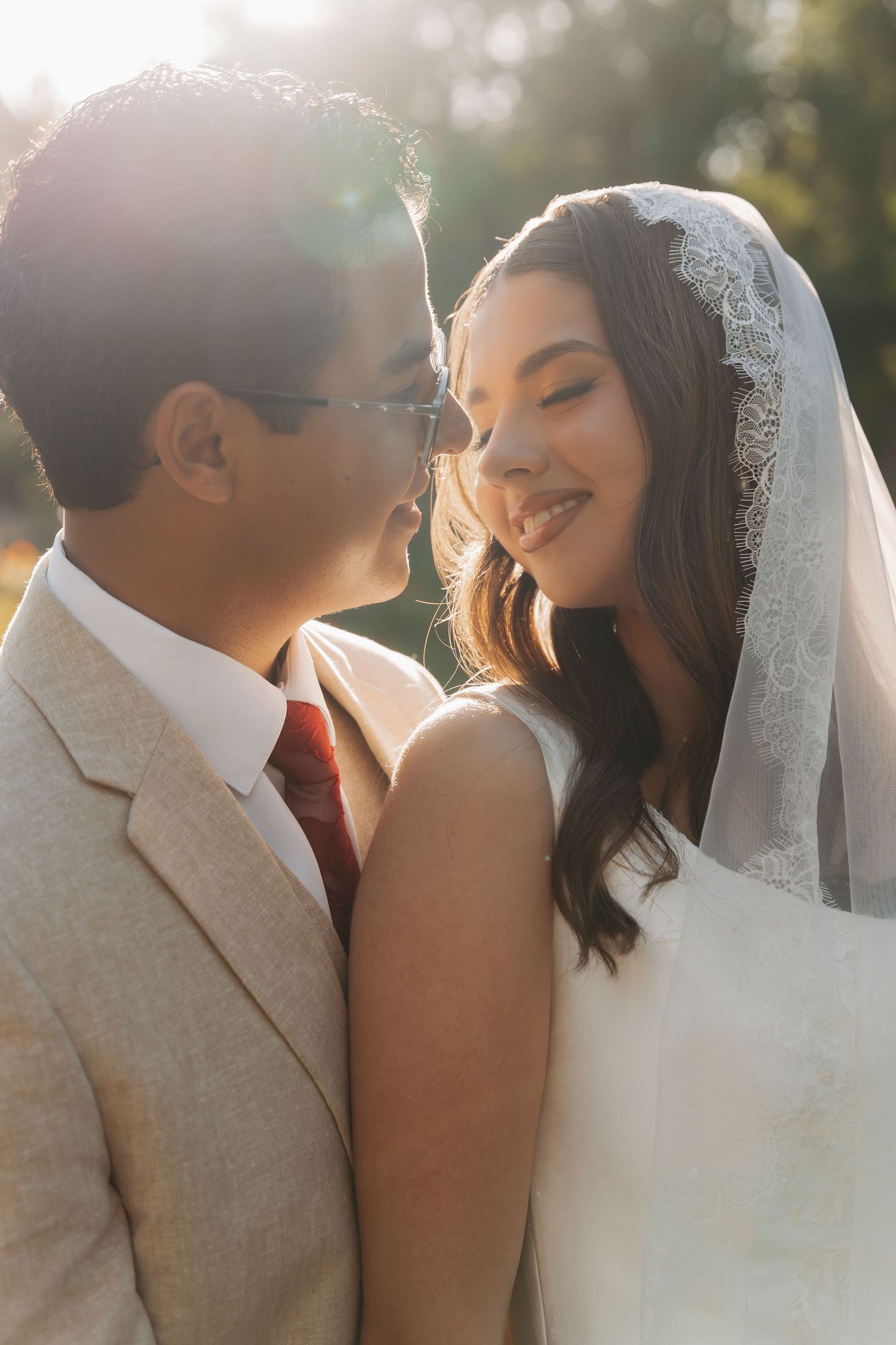 A bride in a lace veil and groom in a tan suit share a romantic moment outdoors in warm golden sunlight.