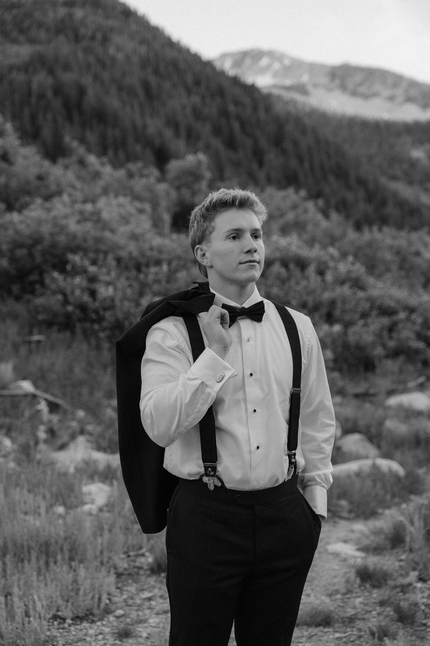 Young man in tuxedo shirt, bow tie, and suspenders holds jacket over shoulder outdoors with mountain backdrop.
