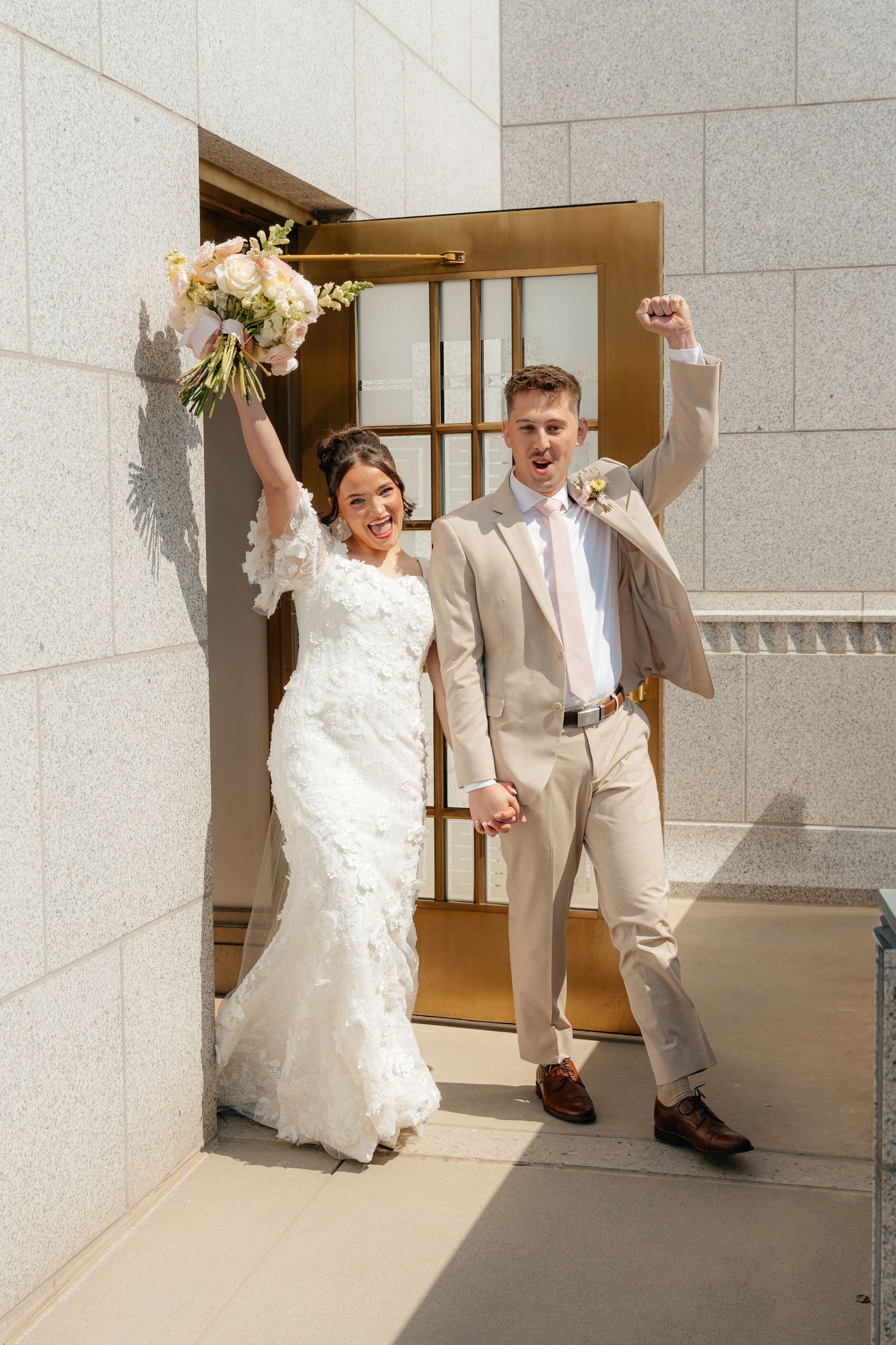 Joyful newlyweds exit wedding venue, bride in lace gown holding bouquet, groom in tan suit, both raising arms in celebration.