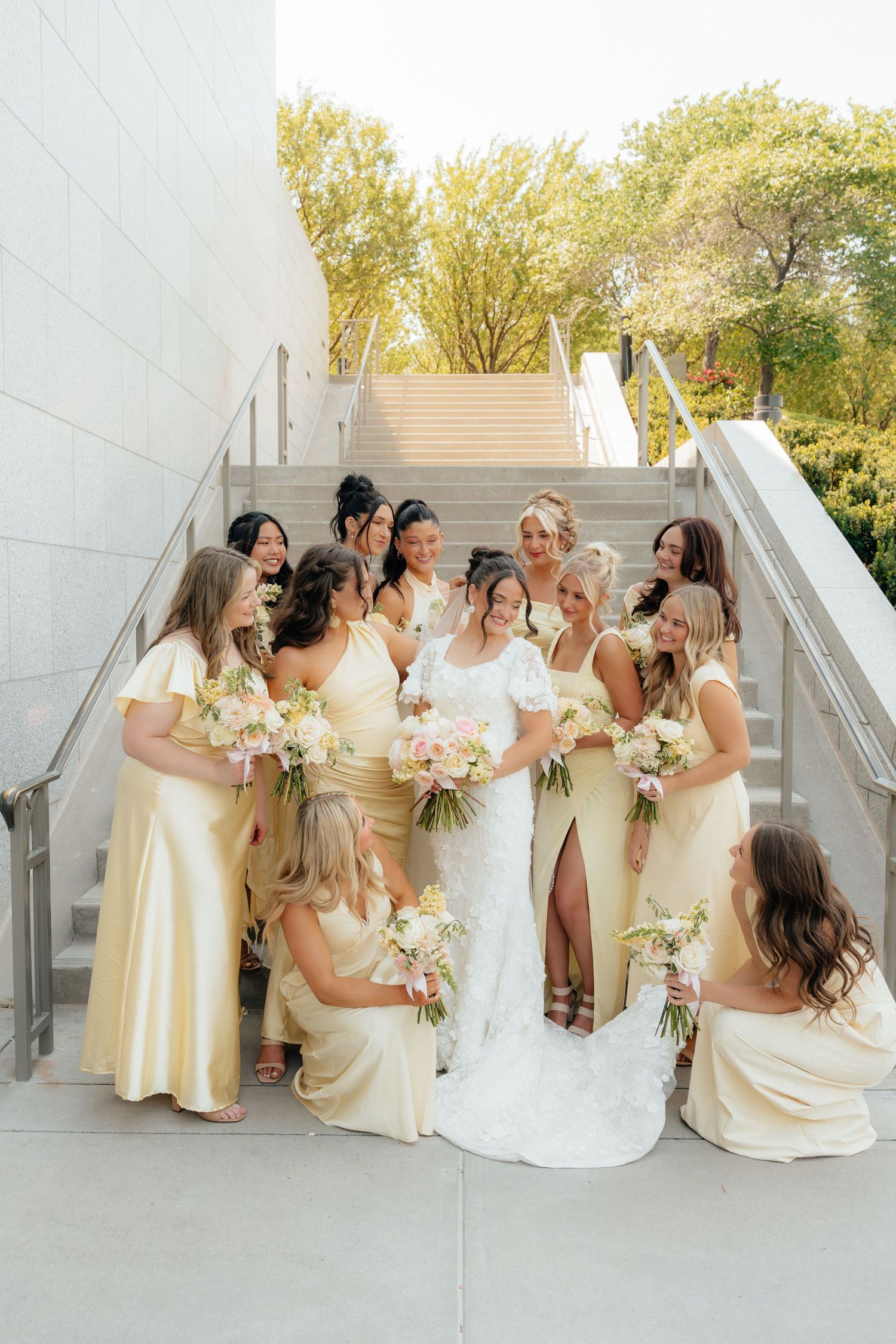 Bride in white lace gown surrounded by bridesmaids in yellow satin dresses holding floral bouquets on outdoor staircase.