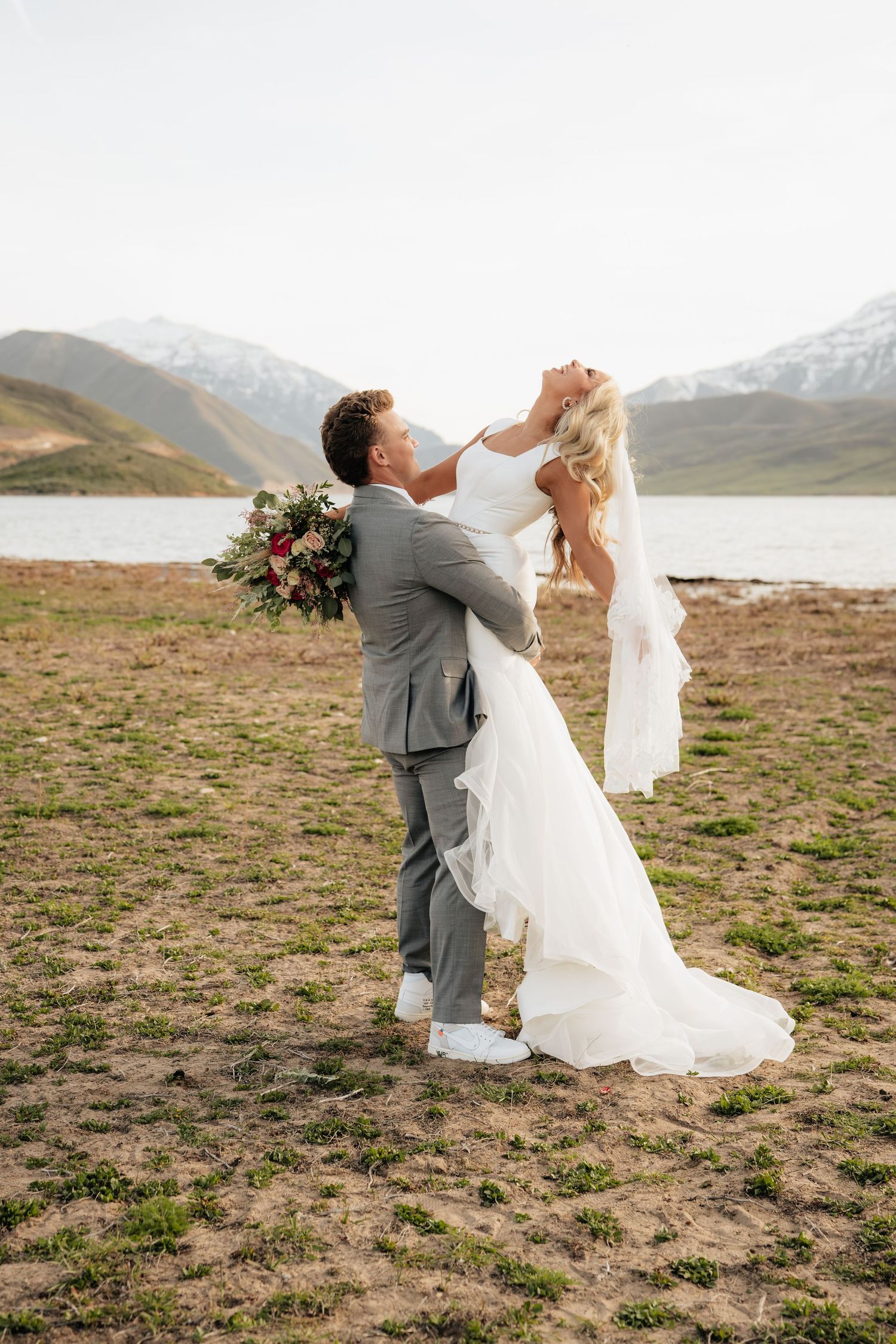 Groom lifts bride in white gown near mountain lake, she holds bouquet, snow-capped peaks in background.