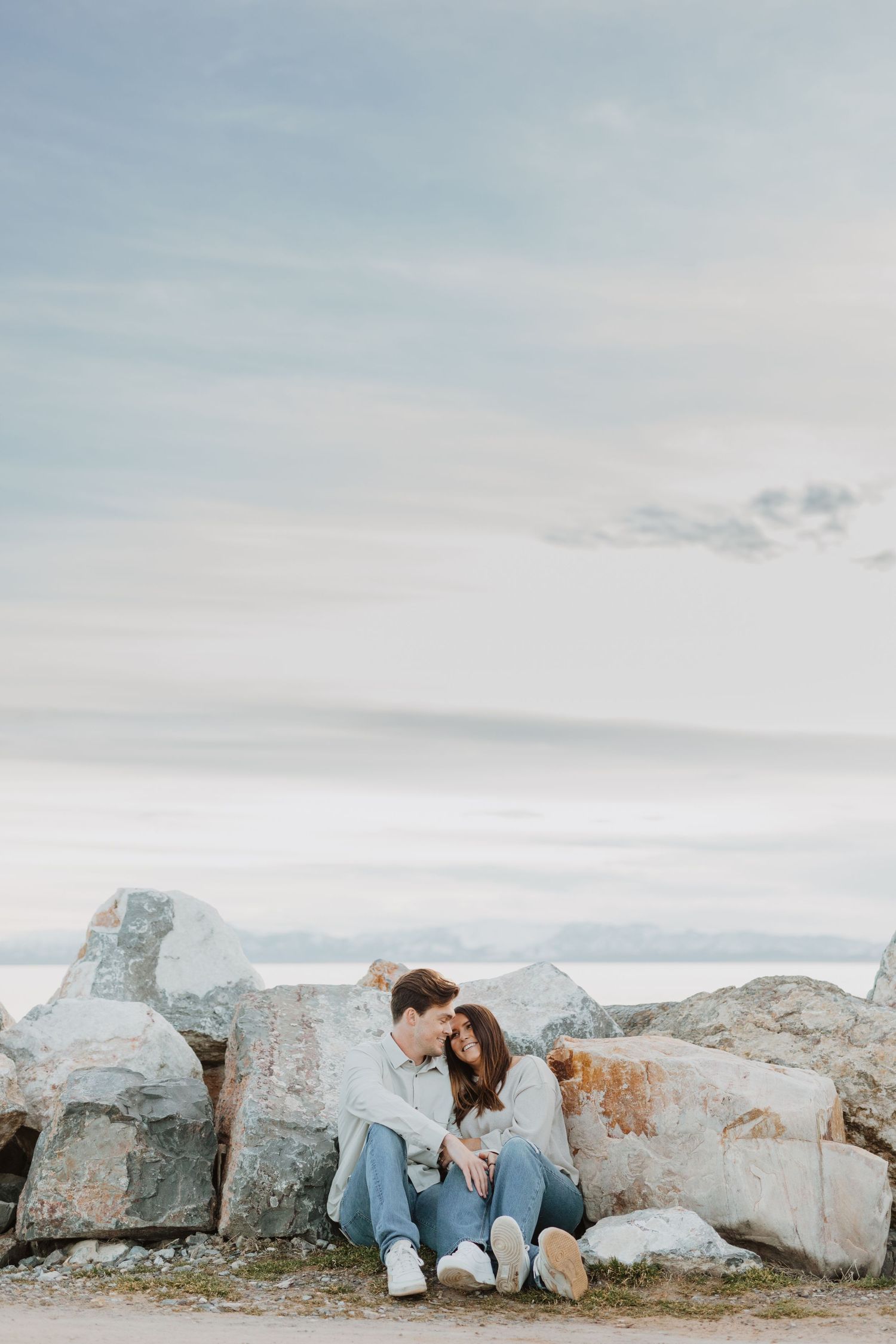 A casual couple sits together on coastal rocks with a misty ocean backdrop.