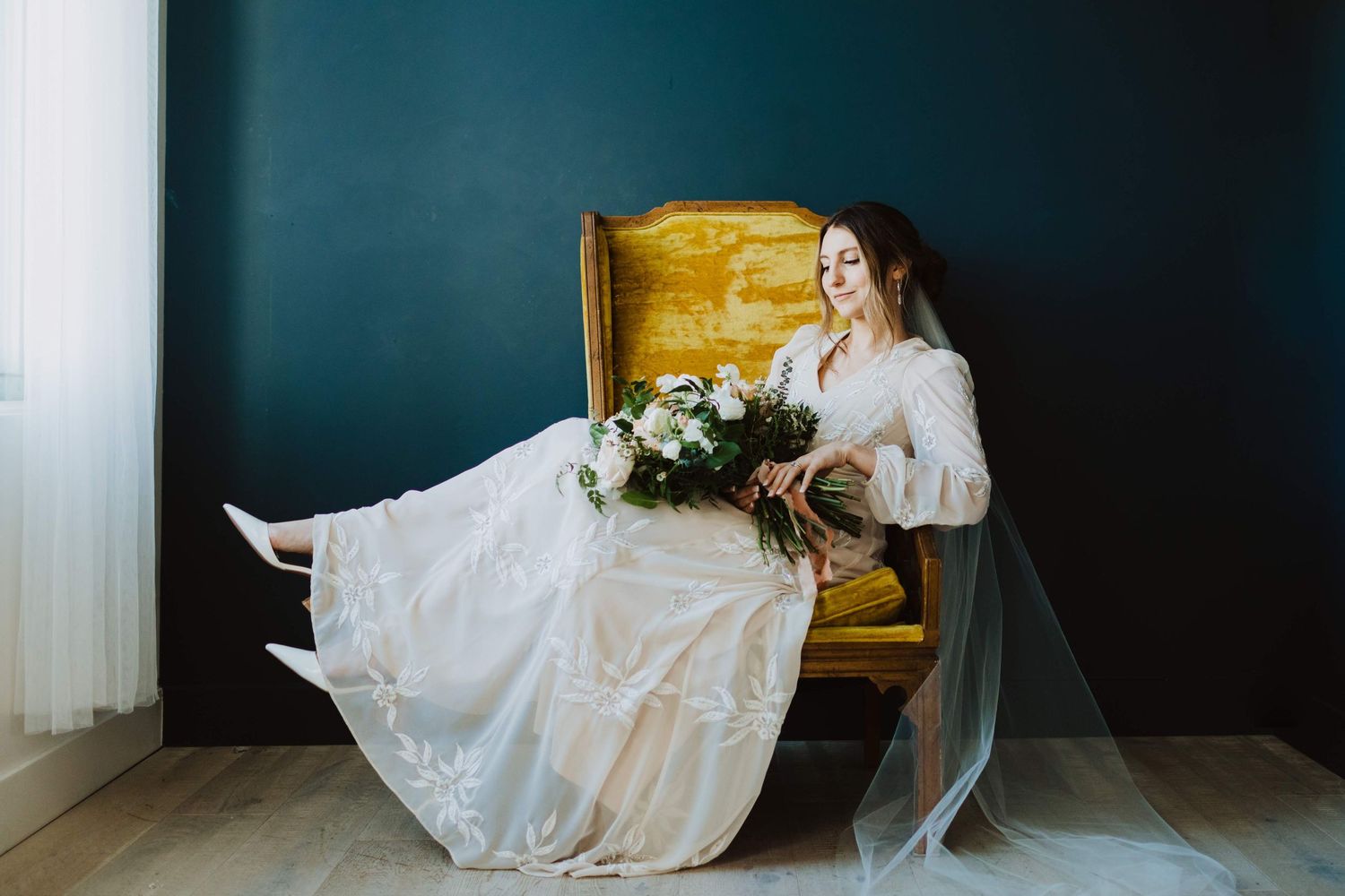 A person in a wedding dress sits elegantly on a wooden chair against a dark teal wall.