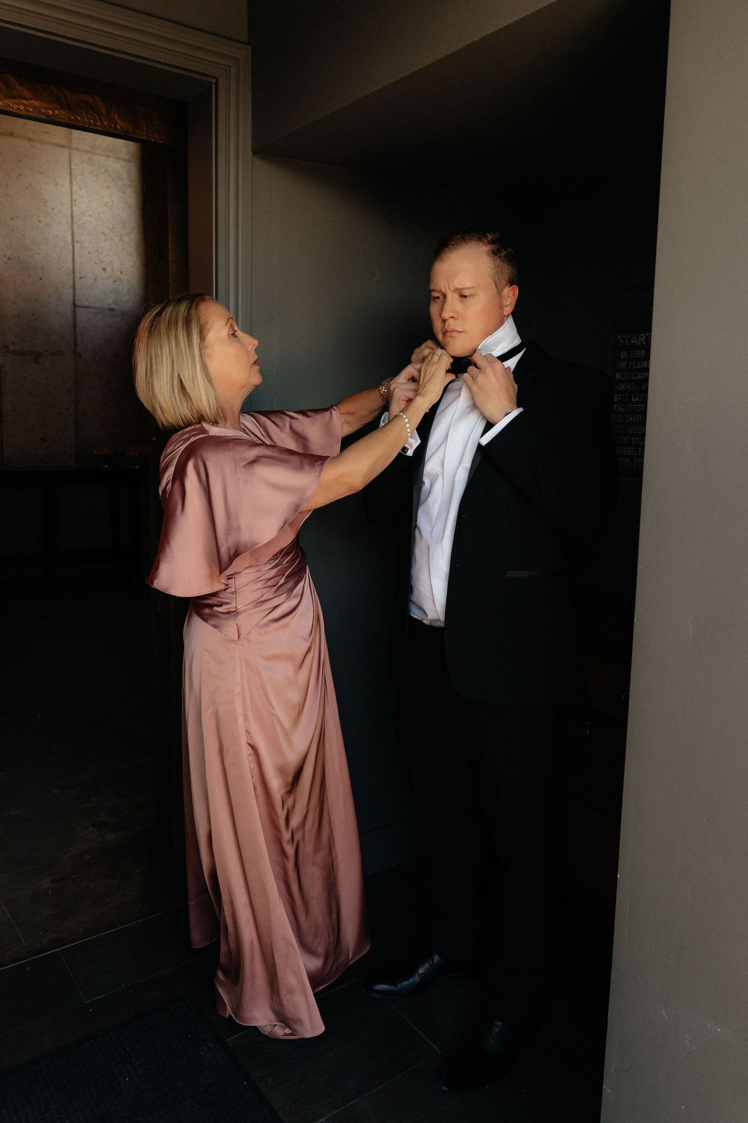 Woman in pink satin gown adjusting bow tie of man in black tuxedo before formal event.