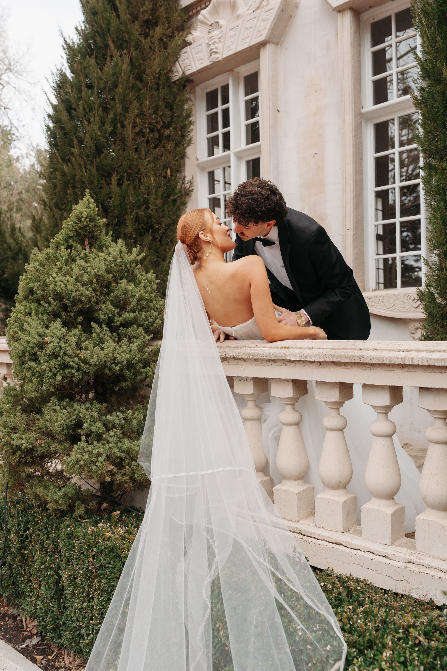 Wedding couple sharing intimate moment on ornate stone balustrade of elegant mansion.