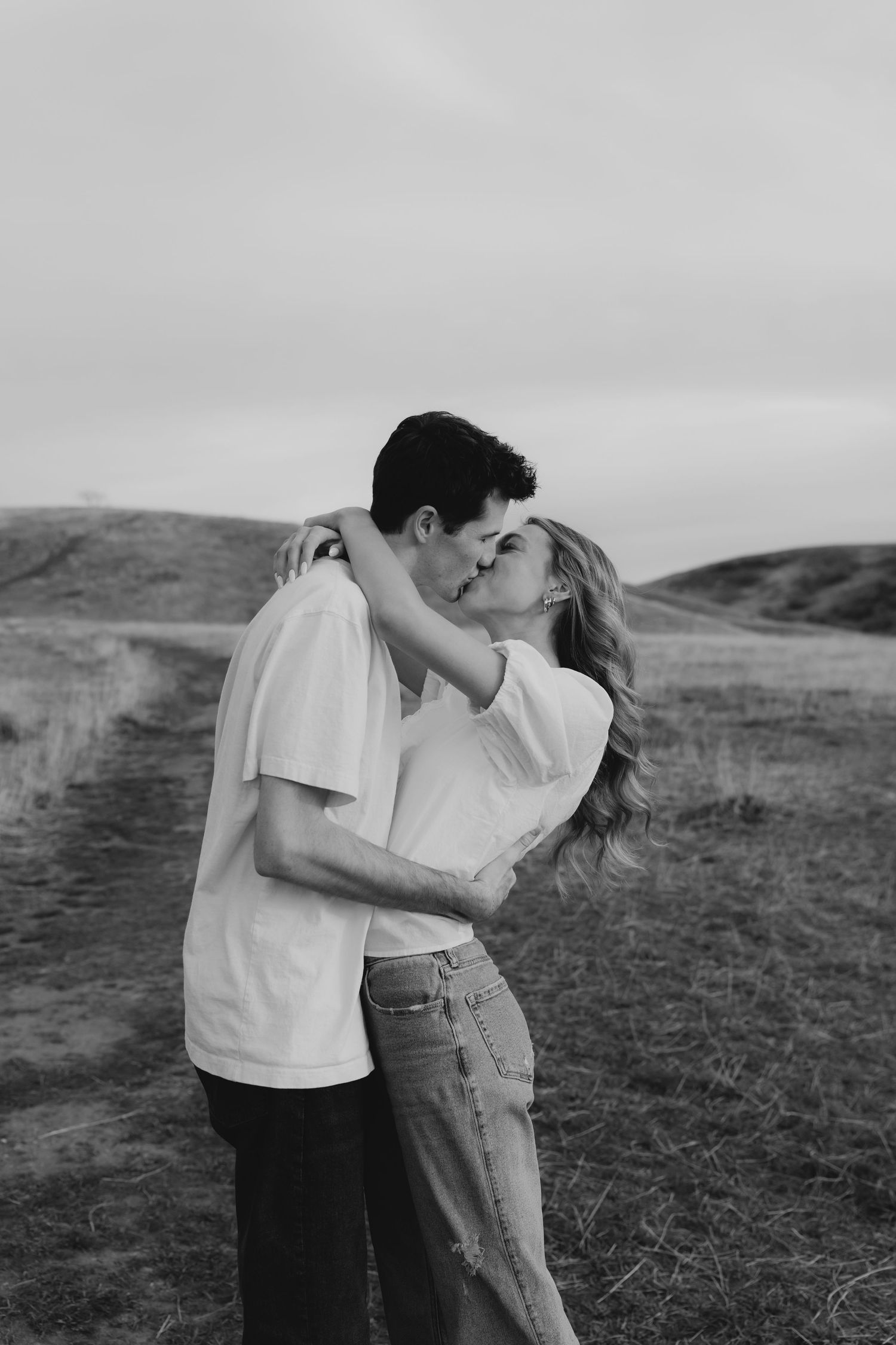 A couple shares a romantic kiss in a black and white photo set against rolling hills and an open field.